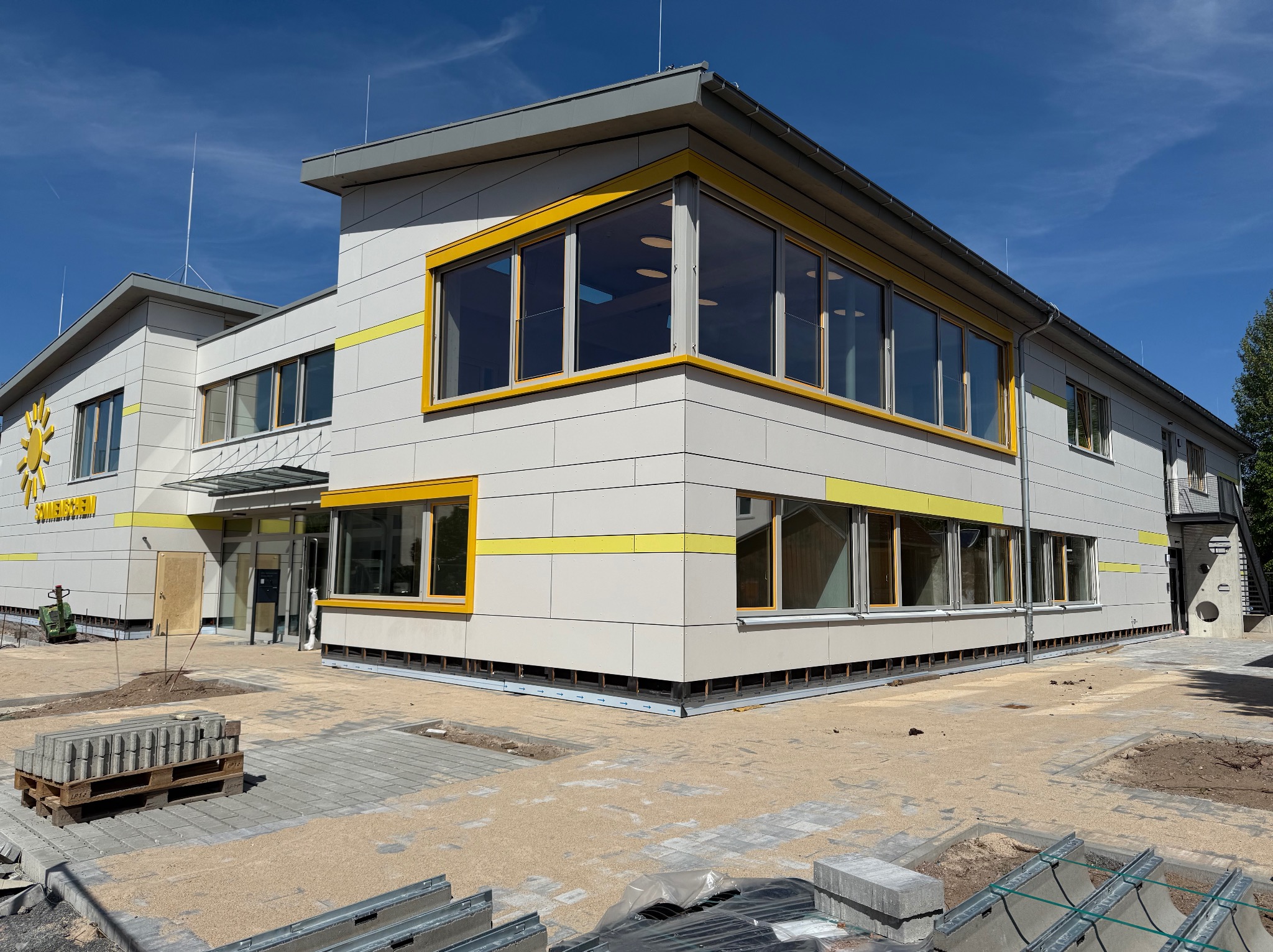 Nowoczesny budynek z jasną elewacją i żółtymi akcentami wokół okien, w trakcie prac wykończeniowych na zewnątrz. Widoczne materiały budowlane i maszyny.