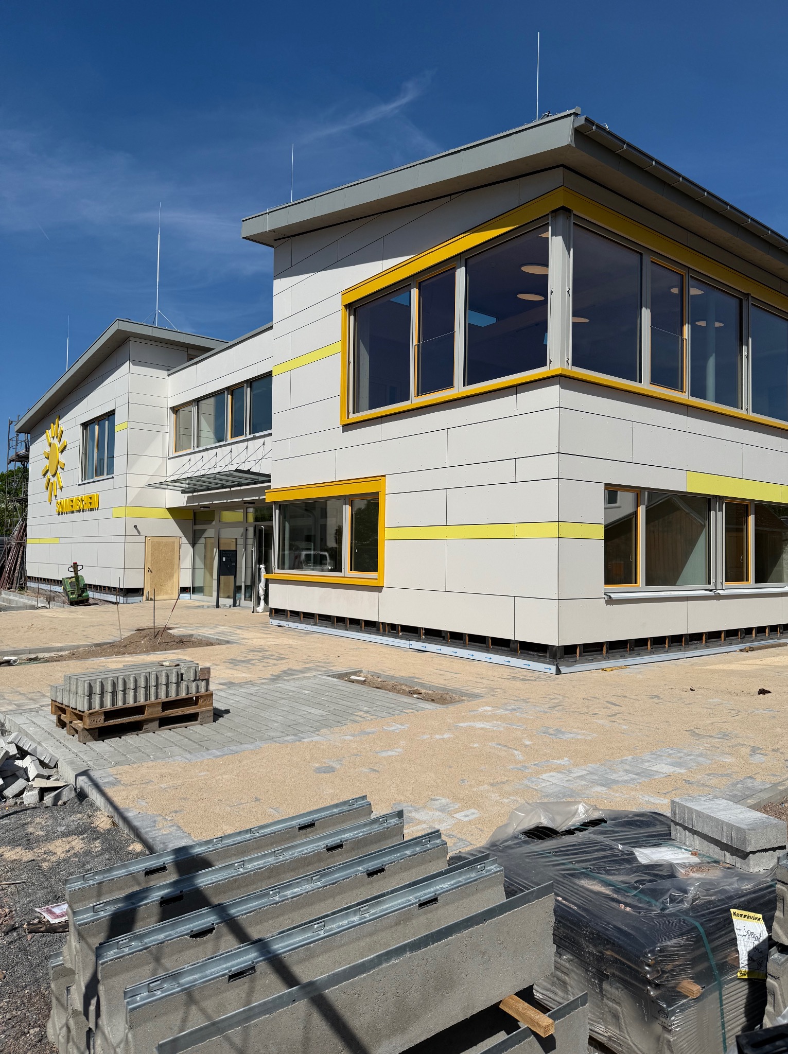 Budynek z jasną elewacją i żółtymi akcentami, w trakcie prac wykończeniowych. Widoczne płyty elewacyjne, kostka brukowa i materiały budowlane.