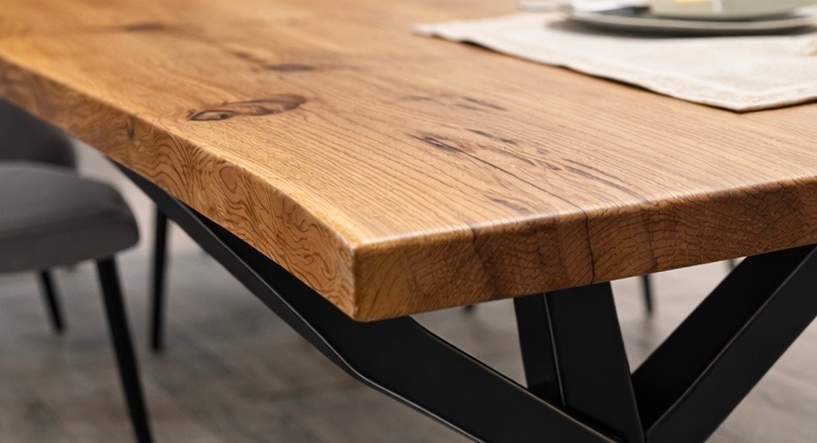 Blat drewniany stołu z wyraźnym usłojeniem i czarną, metalową podstawą w kształcie litery X. Na blacie widoczny fragment nakrycia stołu.