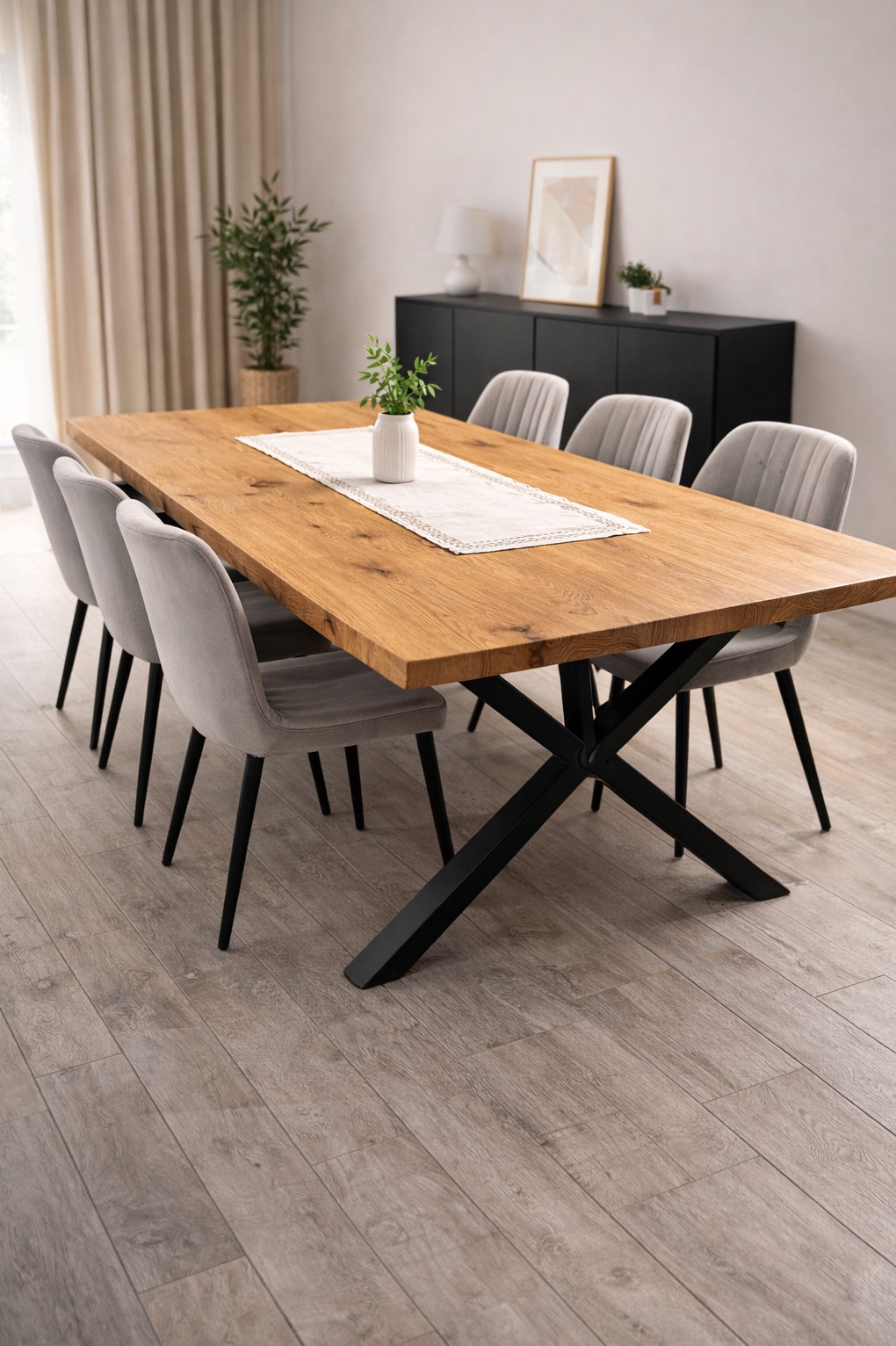 Stół z blatem z jasnego drewna i czarną, metalową podstawą w kształcie litery X, otoczony szarymi krzesłami. Na stole dekoracyjny bieżnik i wazon z zielenią.