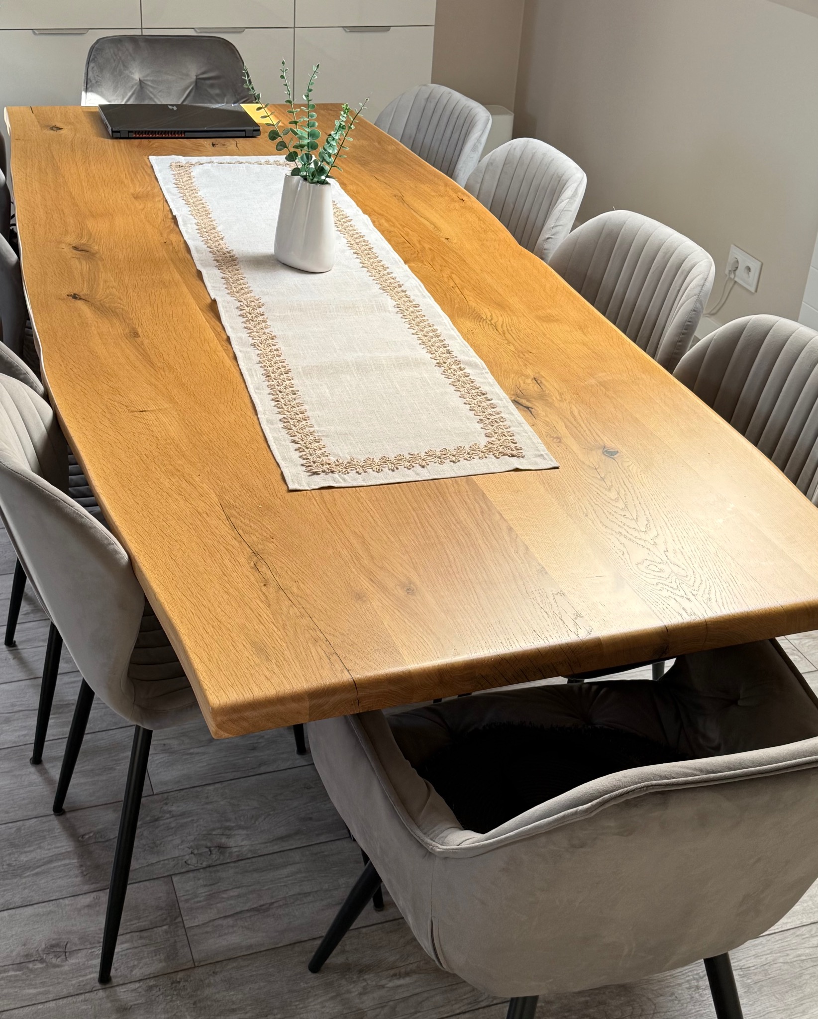 Stół z blatem z litego drewna dębowego, otoczony krzesłami z szarej tapicerki. Na blacie dekoracyjny bieżnik i wazon z zielonymi gałązkami. Widoczny laptop.