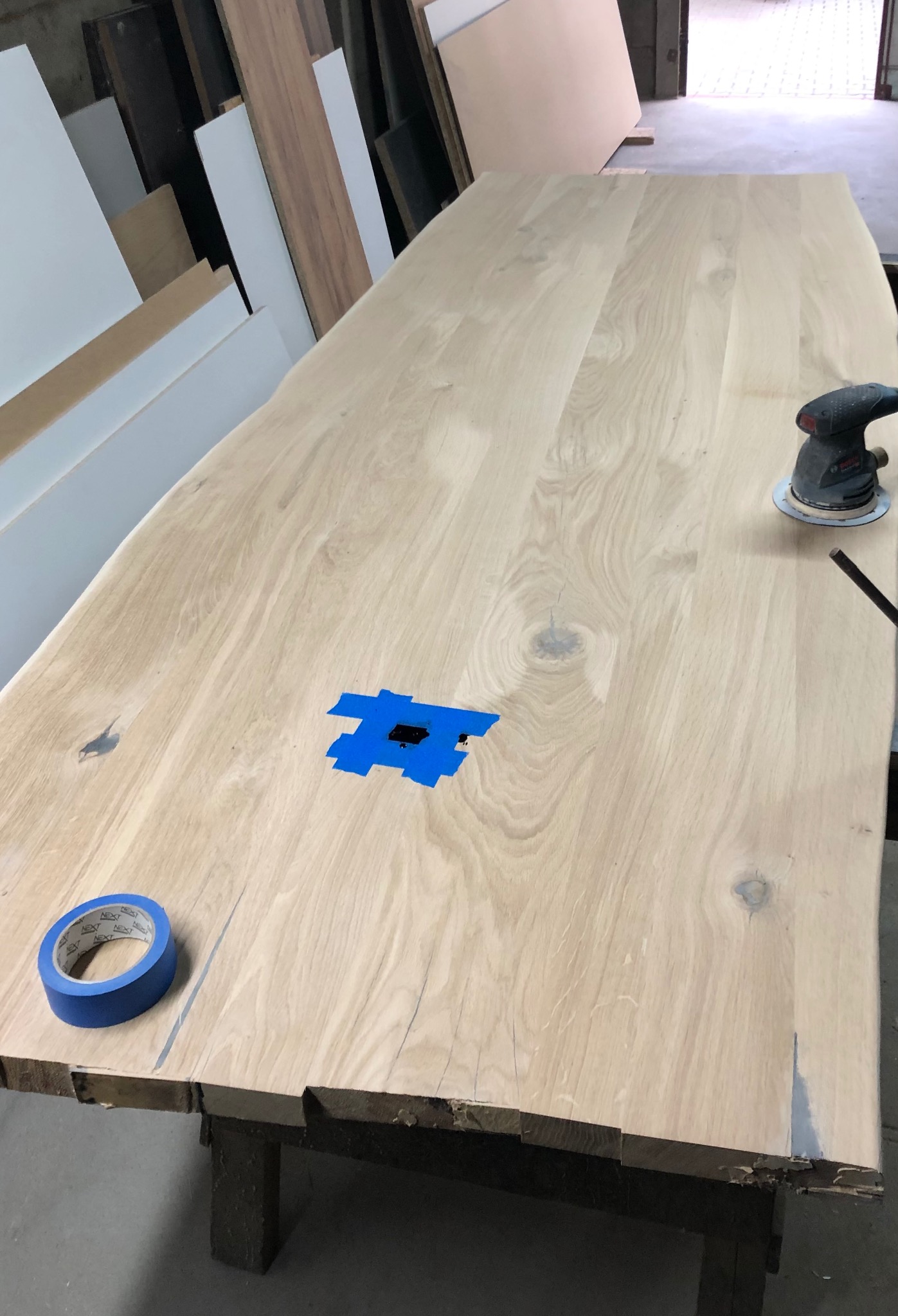 Blat drewniany w trakcie obróbki: szlifowanie z użyciem szlifierki oscylacyjnej, zabezpieczony taśmą malarską w miejscu uszkodzeń. Widoczne słoje drewna.