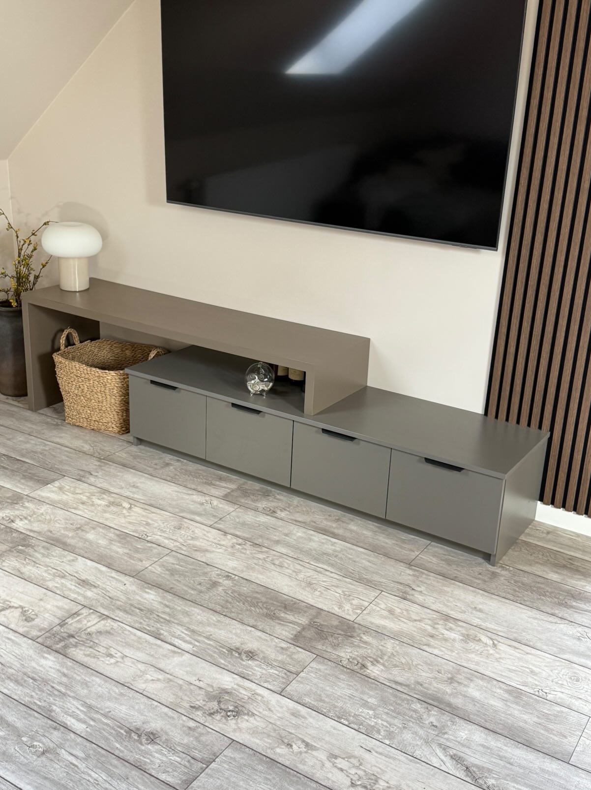 Szara szafka RTV na wymiar z szufladami i półką, dopasowana do wnętrza. Realizacja z Wrześni. Na blacie dekoracje, obok lamelki ścienne i kosz.