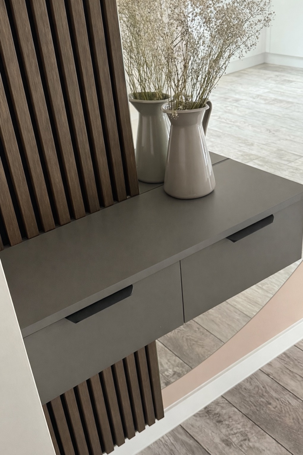 Szara szafka z dwiema szufladami i dekoracyjnym panelem z drewnianych listewek. Na blacie dwa wazony z suszonymi kwiatami. Minimalistyczny design.
