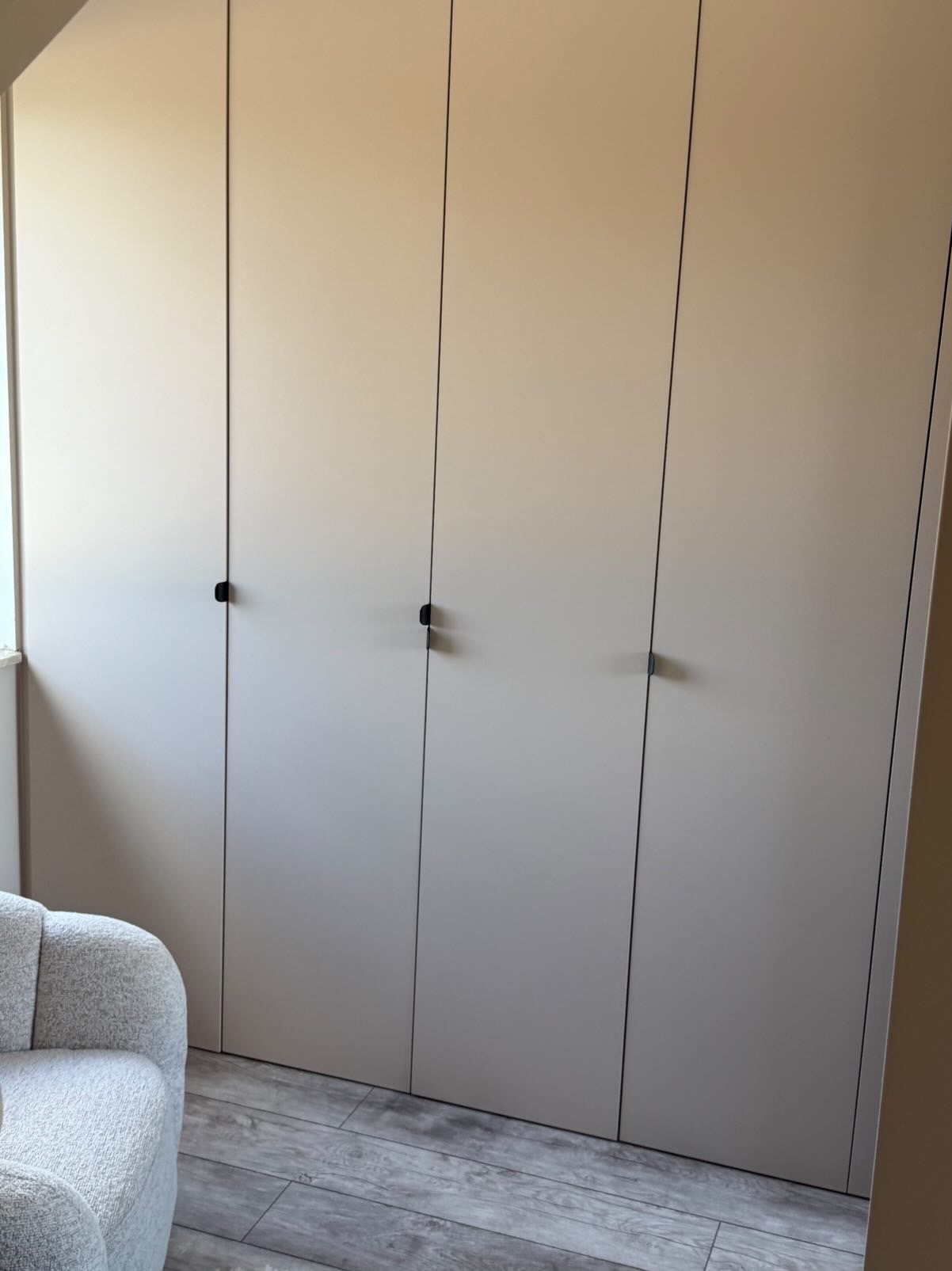 Beżowa szafa wnękowa z minimalistycznymi, czarnymi uchwytami, dopasowana do wnętrza z szarą podłogą i fotel w stylu bouclé. Nowoczesny design.