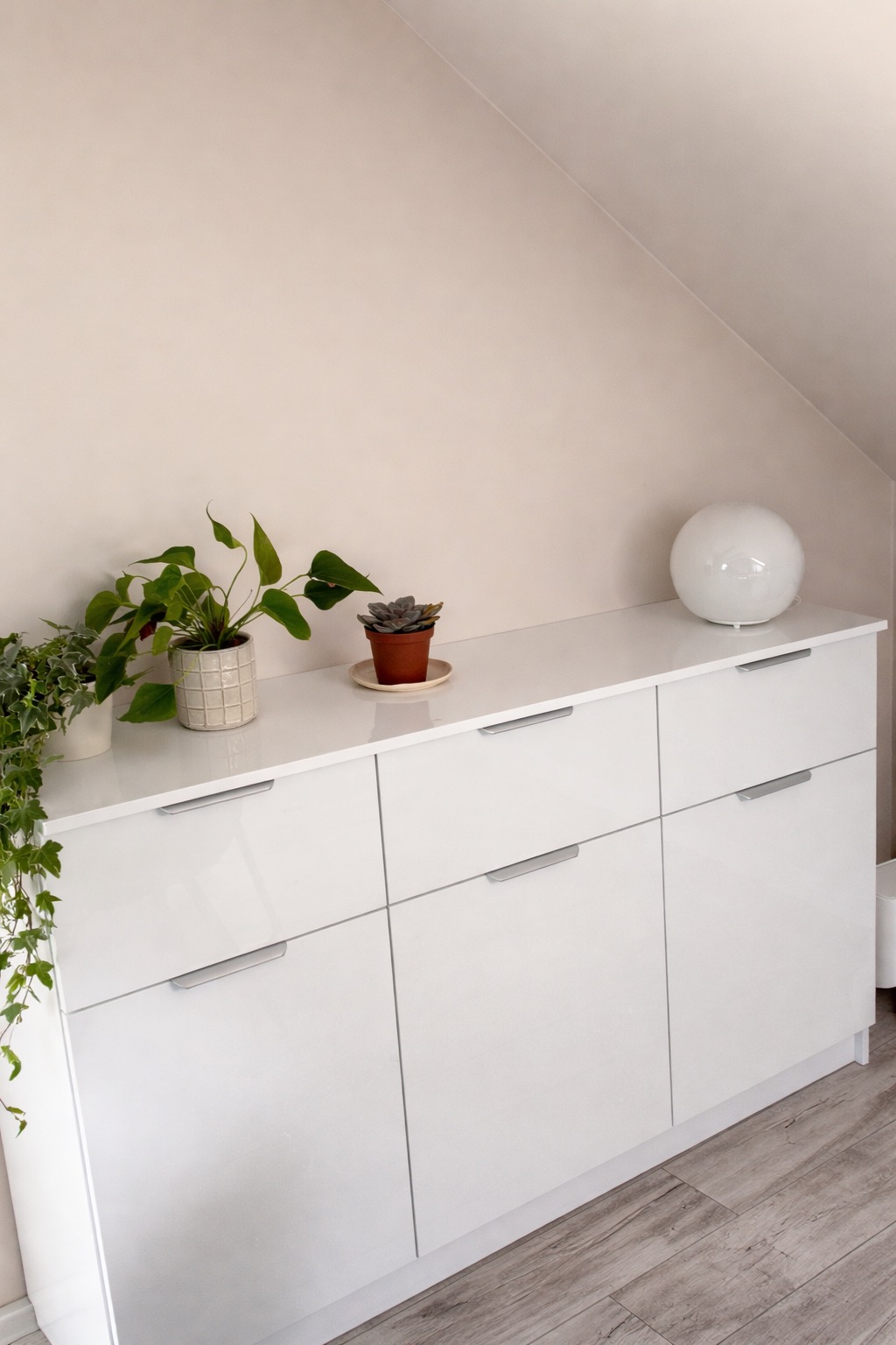 Biała, lakierowana komoda na wymiar we Wrześni, z sześcioma szufladami z minimalistycznymi uchwytami. Na blacie rośliny doniczkowe i okrągła lampa. Ustawiona na poddaszu.