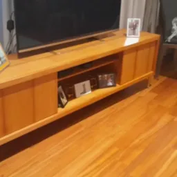 Drewniana szafka RTV z widocznymi półkami i ramkami ze zdjęciami. Na szafce stoi telewizor, obok krzesło z folią ochronną. Parkiet na podłodze.