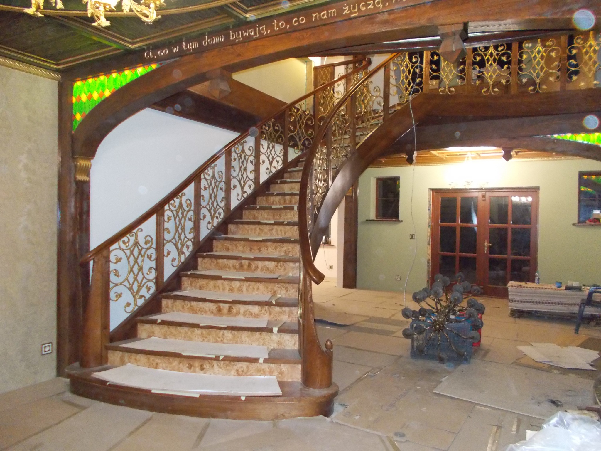Drewniane schody z dekoracyjną, złotą balustradą w bogato zdobionym wnętrzu. Widoczne zabezpieczenia na stopniach podczas prac wykończeniowych.