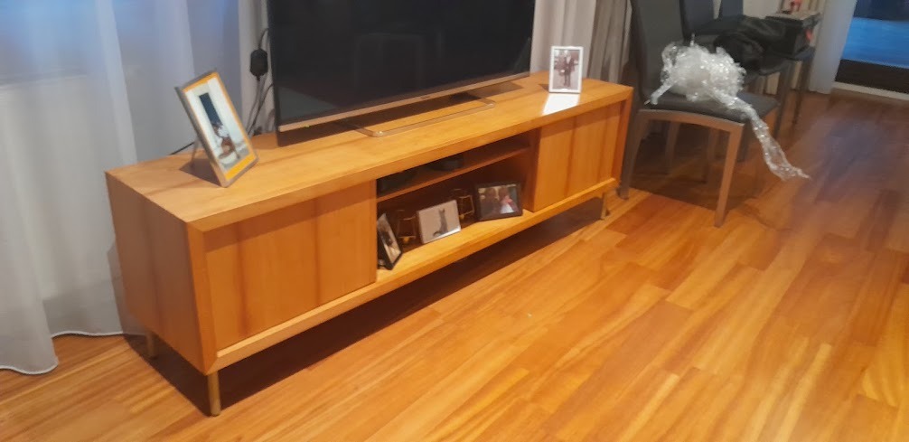 Drewniana szafka RTV z widocznymi półkami i ramkami ze zdjęciami. Na szafce stoi telewizor, obok krzesło z folią ochronną. Parkiet na podłodze.