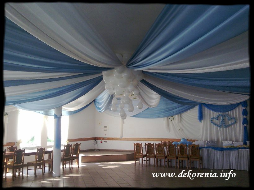 Dekoracja sali weselnej: biało-błękitne drapowania sufitowe, kolumna ozdobiona tkaniną, dekoracja ścienna z sercami, aranżacja stołów dla gości.