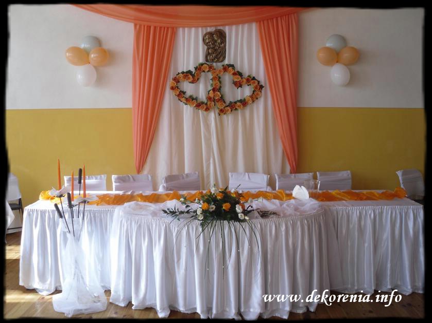 Dekoracja sali okolicznościowej: białe obrusy, pomarańczowe akcenty, kompozycja kwiatowa na stole, dekoracja ścienna z kwiatów w kształcie serc i draperia.