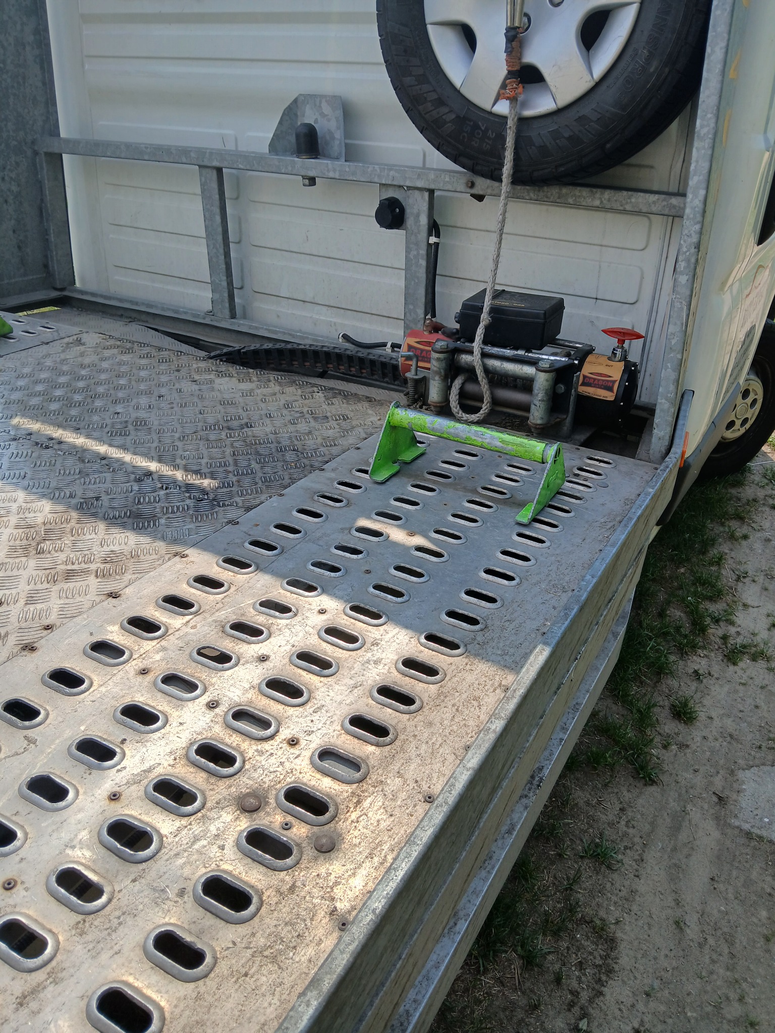 Platforma lawety z wyciągarką i kołem zapasowym. Metalowa powierzchnia z otworami, zielone zabezpieczenia. Widoczne elementy konstrukcyjne i opona.