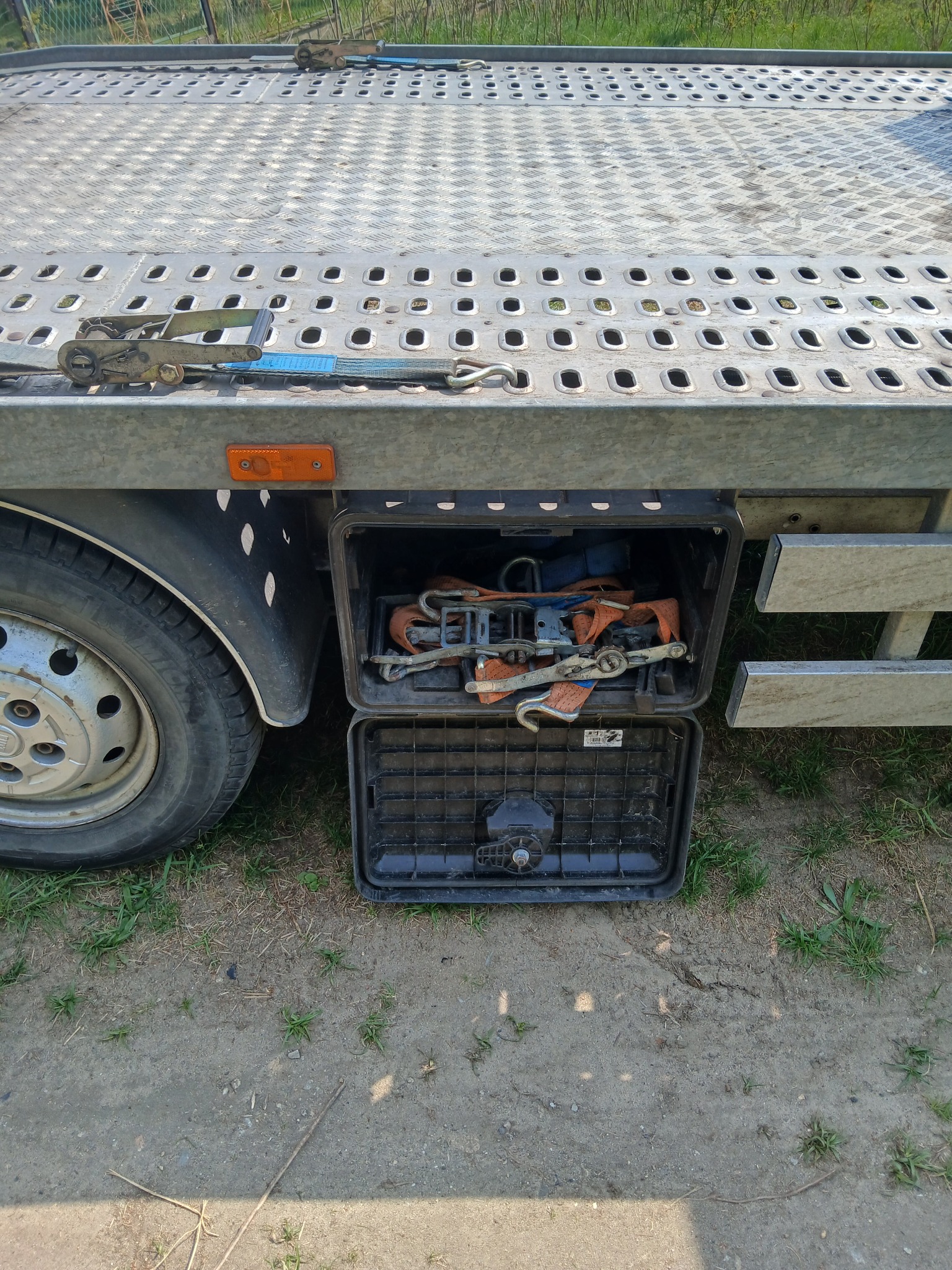 Laweta z otwartym schowkiem, w którym znajdują się pasy transportowe i łańcuchy. Widoczne koło lawety i fragment platformy załadunkowej.