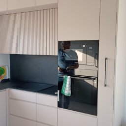 ATP meble - Jasna zabudowa kuchenna z piekarnikiem i mikrofalówką Samsung. Fronty frezowane pionowo, czarny blat i panel nad nim. W odbiciu widoczna osoba.