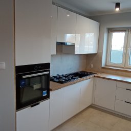 ATP meble - Biała, nowoczesna kuchnia z drewnianym blatem, czarną płytą grzewczą i piekarnikiem w zabudowie. Jasne płytki na podłodze, okno z widokiem na zewnątrz.