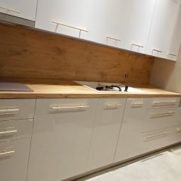 ATP meble - Jasna kuchnia na wymiar z drewnianym blatem i frontami z podłużnymi uchwytami, widoczny fragment płyty grzewczej i zlewu. Na drugim planie osoby.