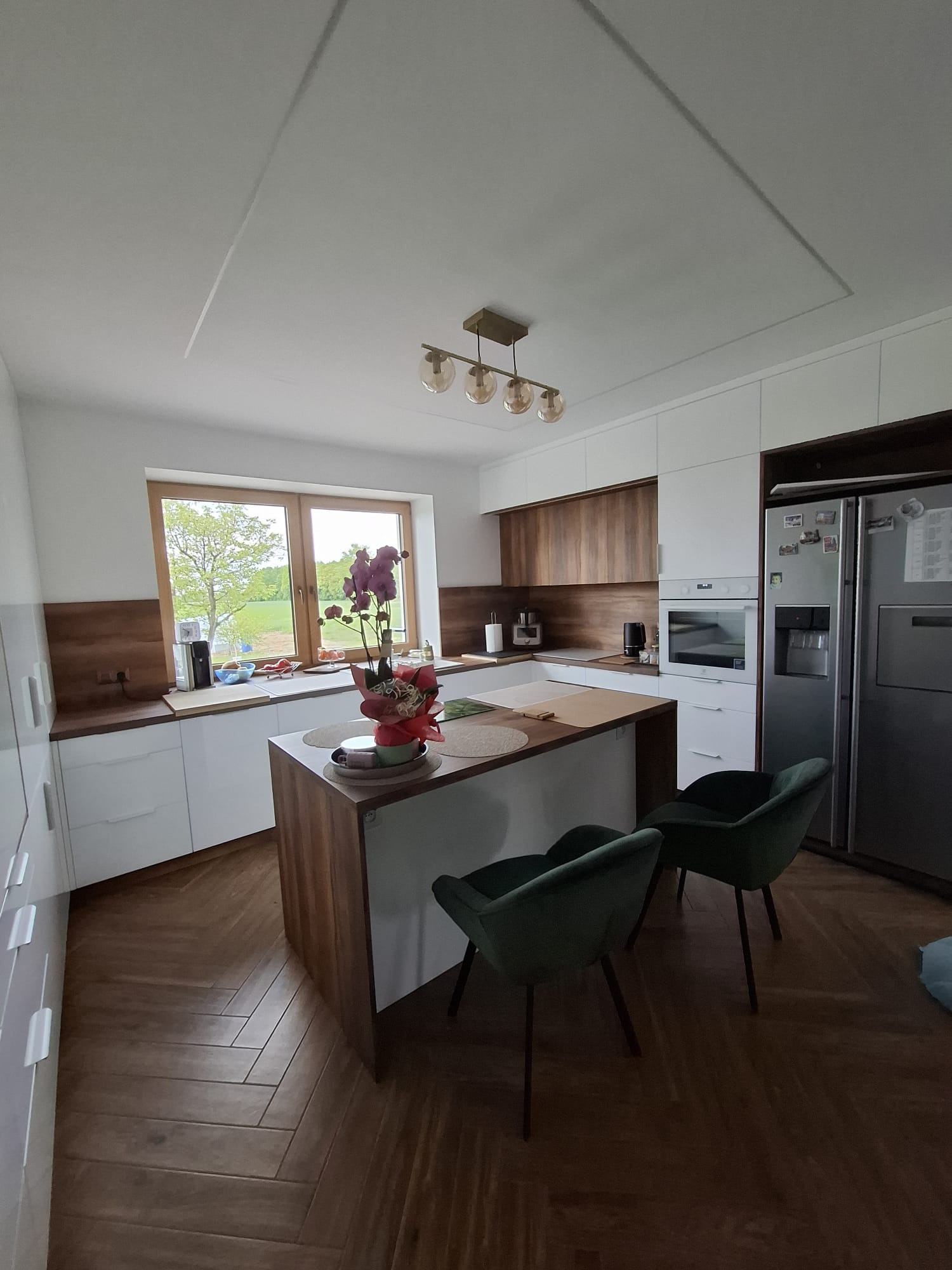 Nowoczesna kuchnia z wyspą, drewnianym blatem i białymi frontami, z zielonymi krzesłami i parkietem w jodełkę. Na wyspie stoi kwiat w doniczce.