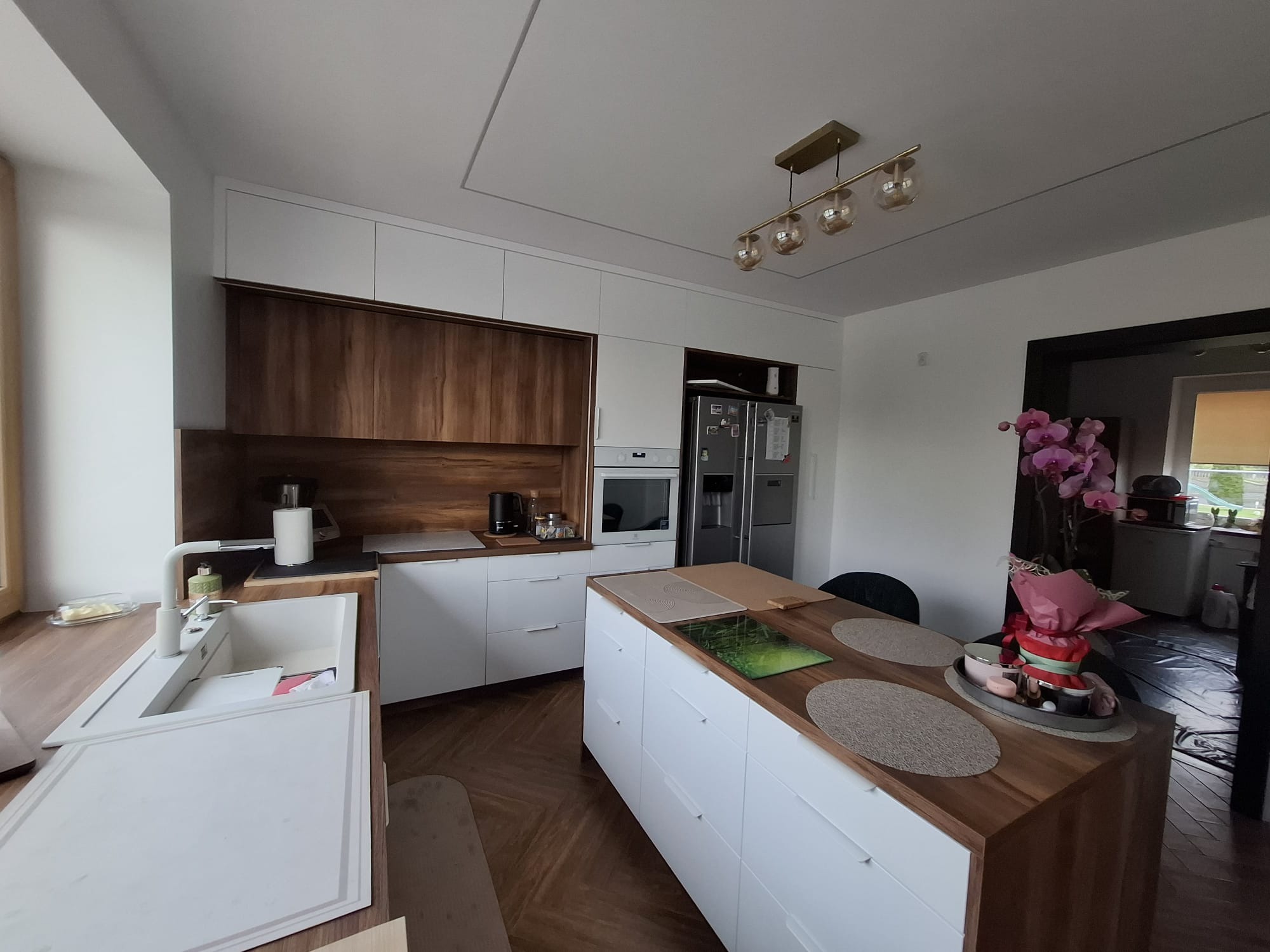 Nowoczesna kuchnia z wyspą: białe fronty, drewniane blaty i szafki górne. Oświetlenie sufitowe i parkiet w jodełkę dodają elegancji. Widoczny zlew i elementy dekoracyjne.