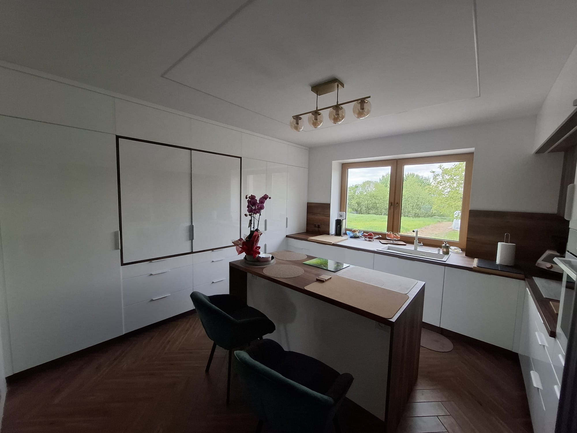 Biała kuchnia z wyspą, ciemne krzesła i parkiet w jodełkę. Okno z widokiem na zieleń. Minimalistyczna lampa sufitowa. Nowoczesny design kuchni na wymiar.