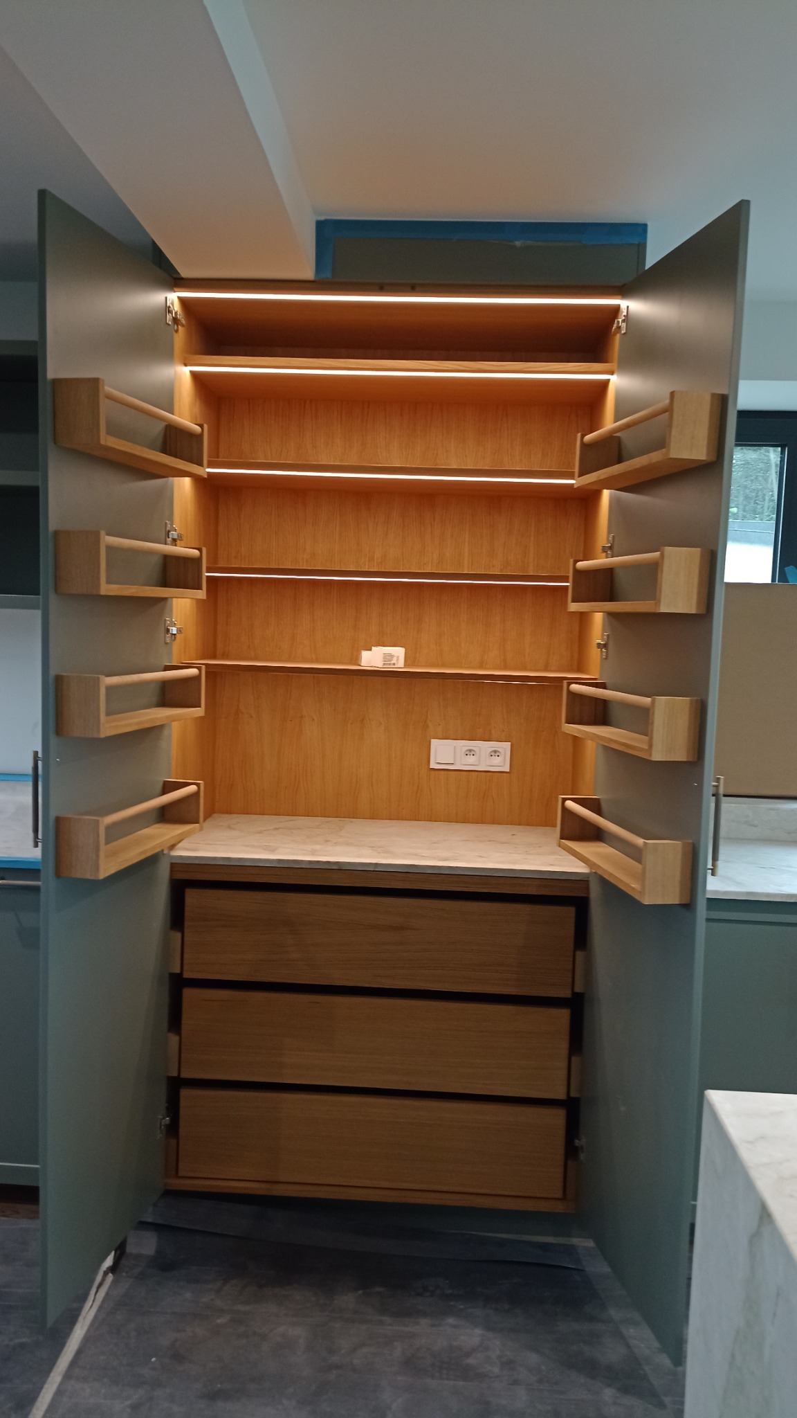 Otwarta, wysoka szafka kuchenna z oświetleniem LED, drewnianymi półkami na przyprawy i trzema szufladami. Fronty w kolorze oliwkowym, wnętrze drewniane.