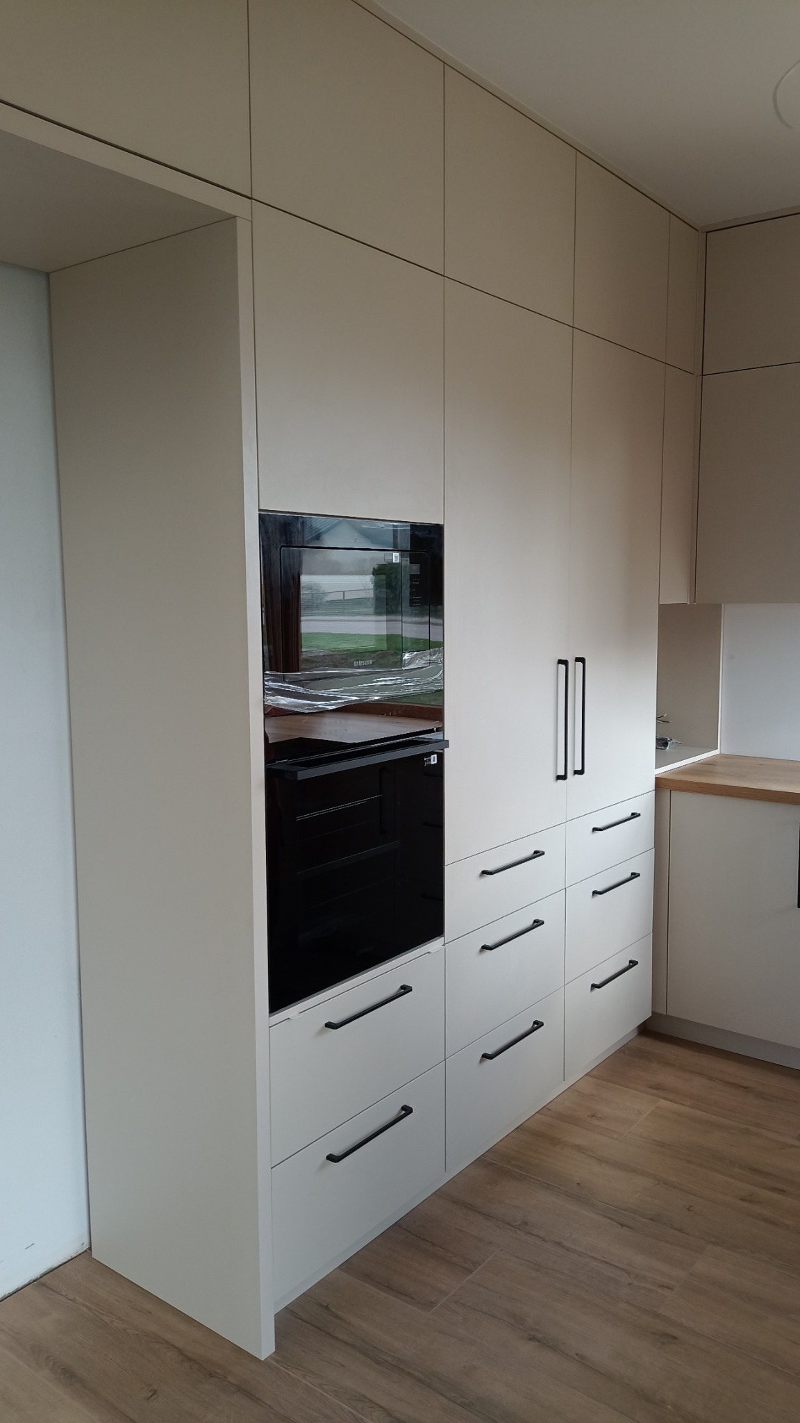 Jasna zabudowa kuchenna z piekarnikiem i szufladami, minimalistyczne czarne uchwyty, drewniana podłoga. Piekarnik zabezpieczony folią ochronną.