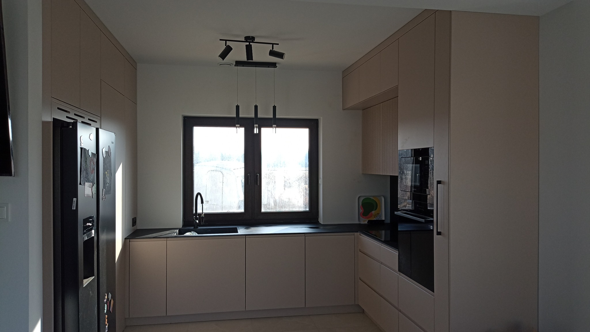 Jasna kuchnia z zabudową po sufit, czarny blat, okno z widokiem na zewnątrz, nowoczesne oświetlenie i czarna lodówka z magnesami.