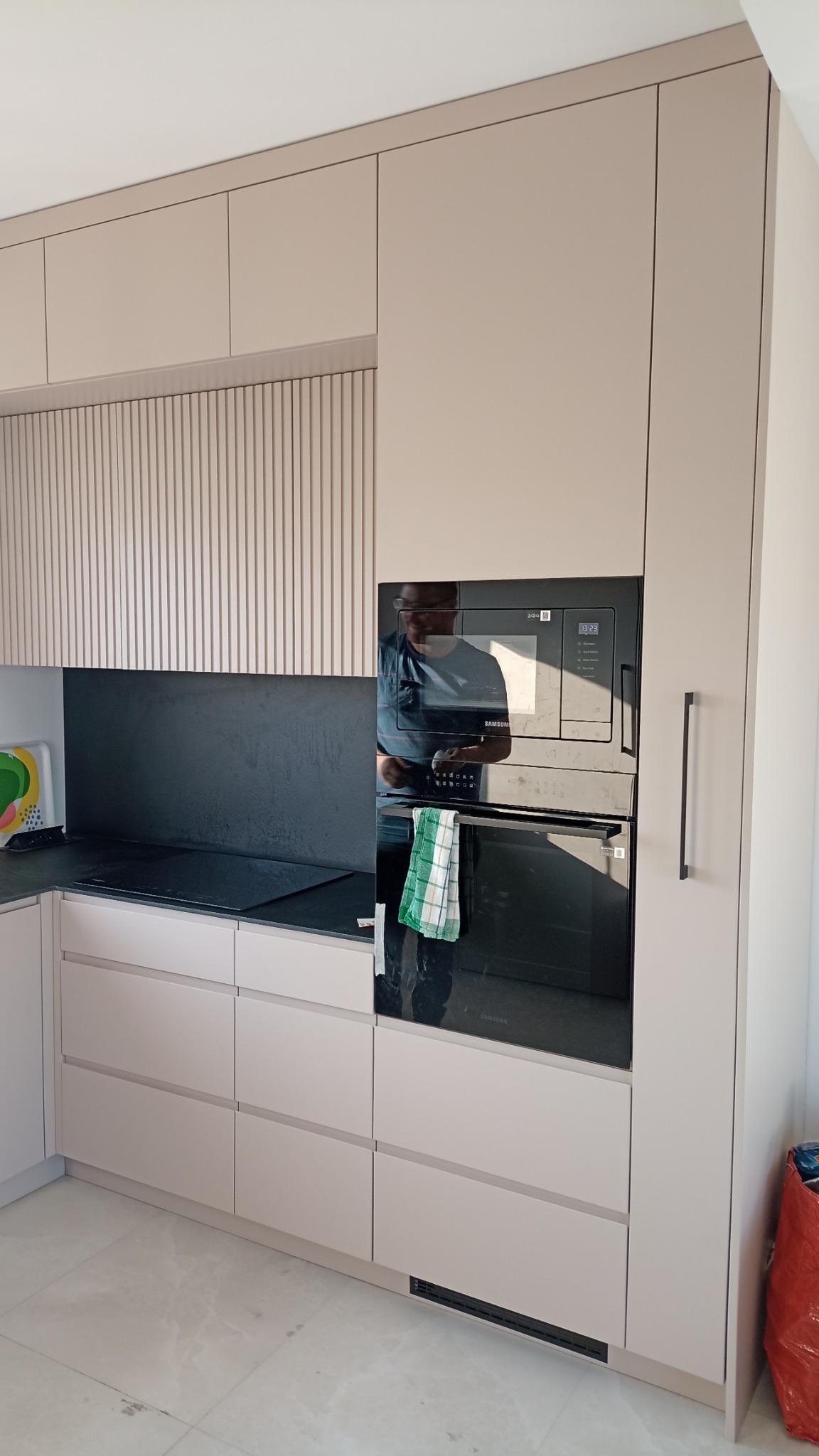 Jasna zabudowa kuchenna z piekarnikiem i mikrofalówką Samsung. Fronty frezowane pionowo, czarny blat i panel nad nim. W odbiciu widoczna osoba.