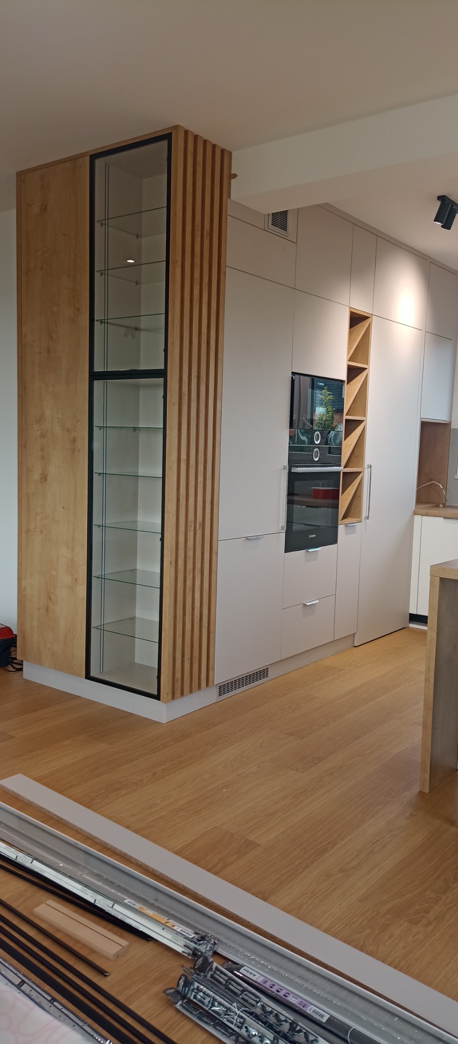 Jasna kuchnia na wymiar z drewnianym akcentem: witryna z półkami i zabudowa piekarnika. Na podłodze leżą elementy montażowe.