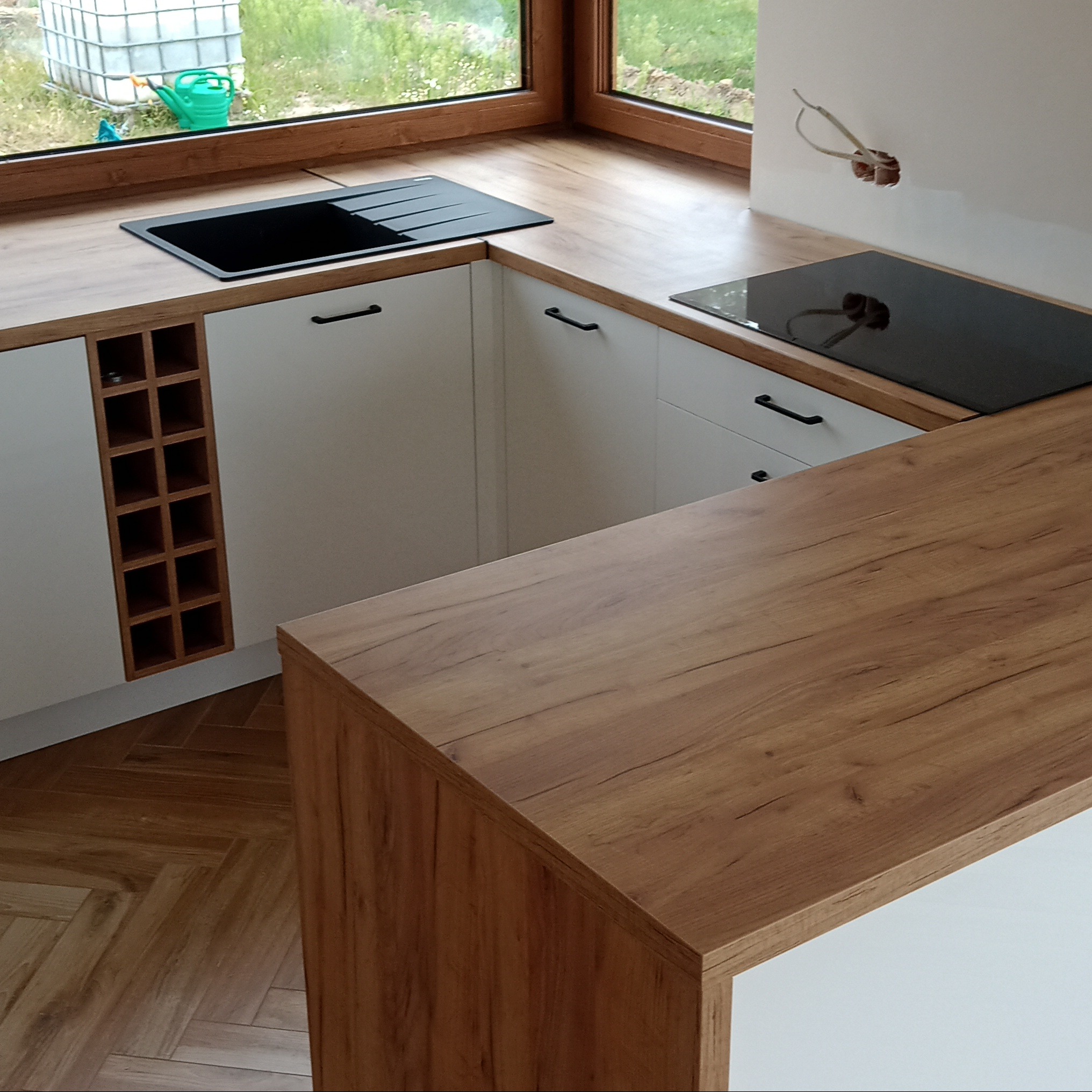 Nowoczesna kuchnia z białymi szafkami, drewnianym blatem i czarnym zlewem. Widoczna półka na wino i płyta indukcyjna. Za oknem zieleń i zbiornik na wodę.