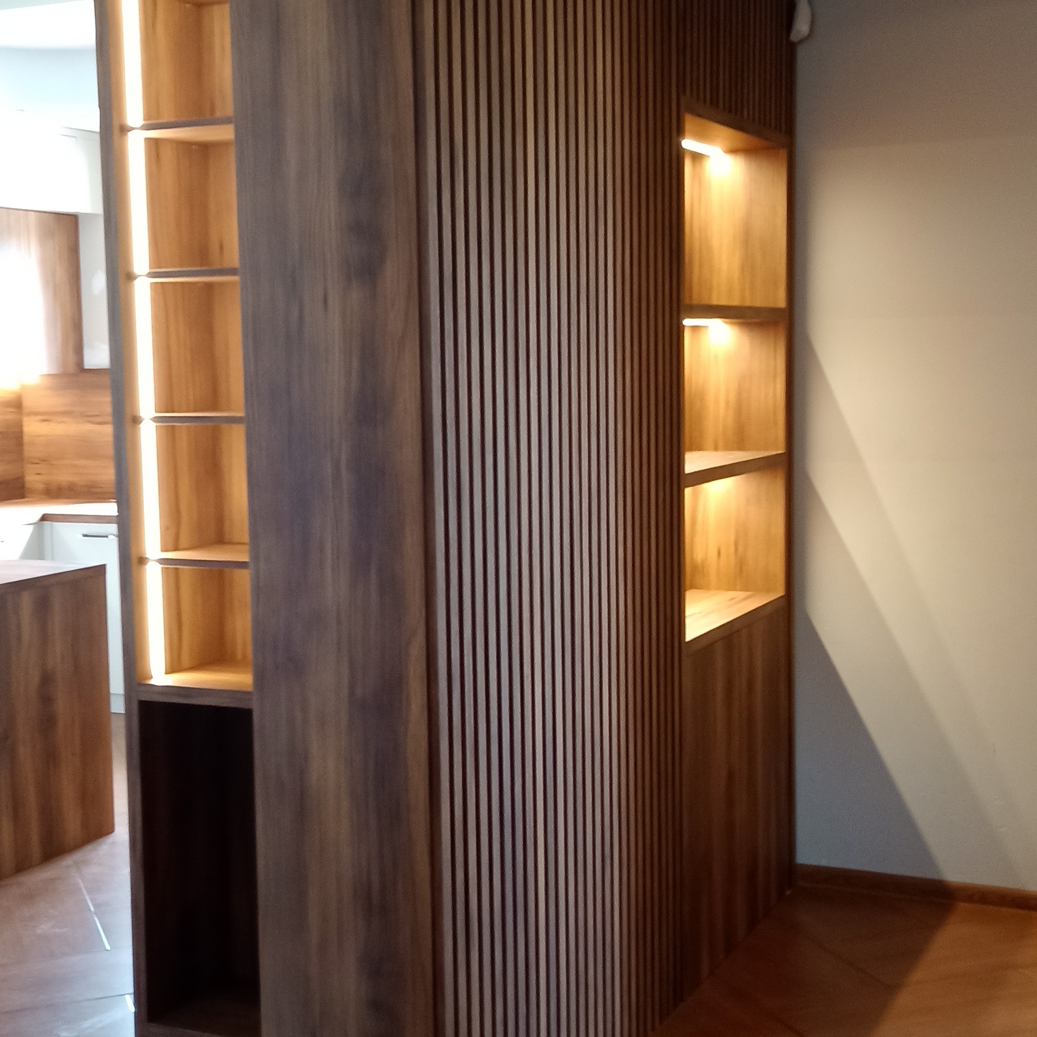 Drewniana zabudowa z pionowymi lamelami i podświetlanymi półkami wnękowymi, widok na kuchnię w tle. Nowoczesny design z ciepłym oświetleniem.