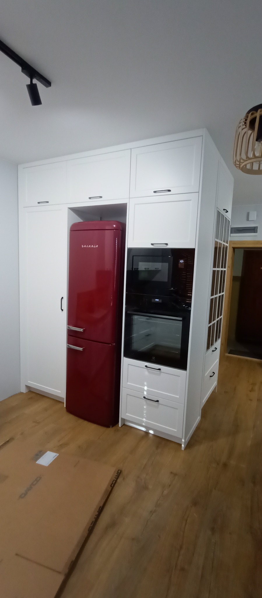 Biała zabudowa kuchenna z czerwonym retro lodówką Gorenje, piekarnikiem i mikrofalówką. Na podłodze leżą kartony. W tle widoczne drzwi.