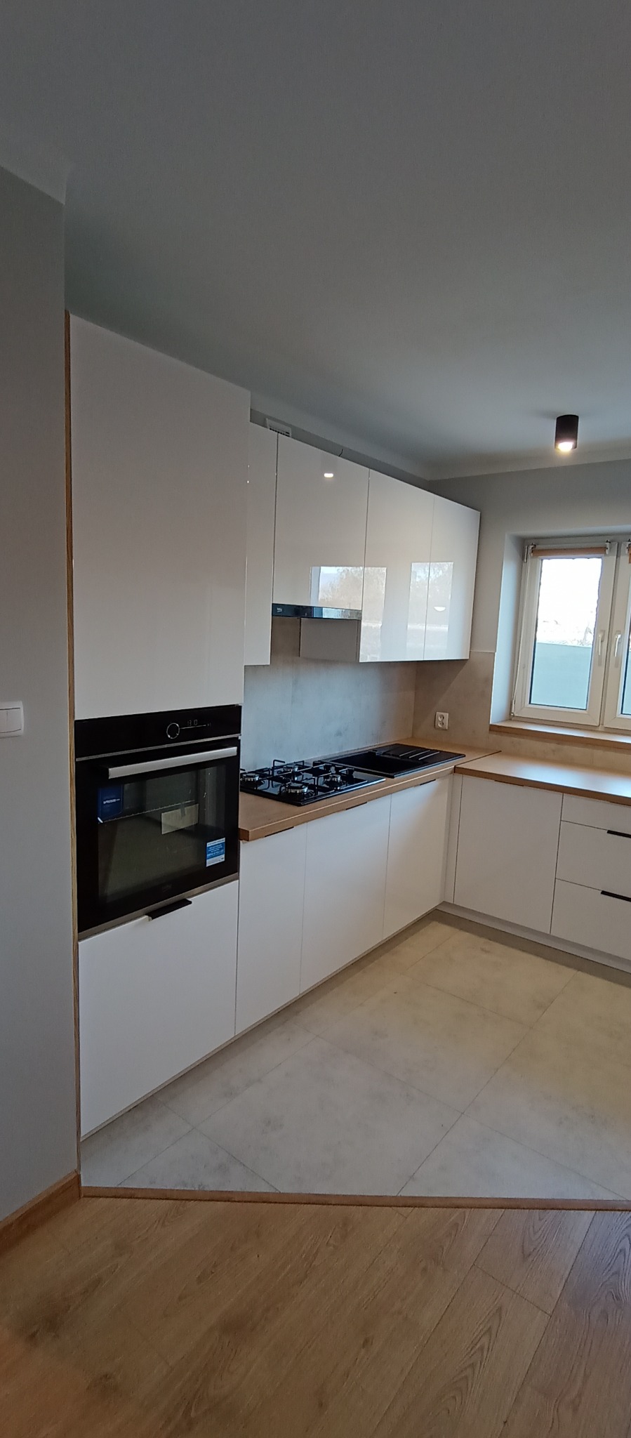 Biała, nowoczesna kuchnia z drewnianym blatem, czarną płytą grzewczą i piekarnikiem w zabudowie. Jasne płytki na podłodze, okno z widokiem na zewnątrz.