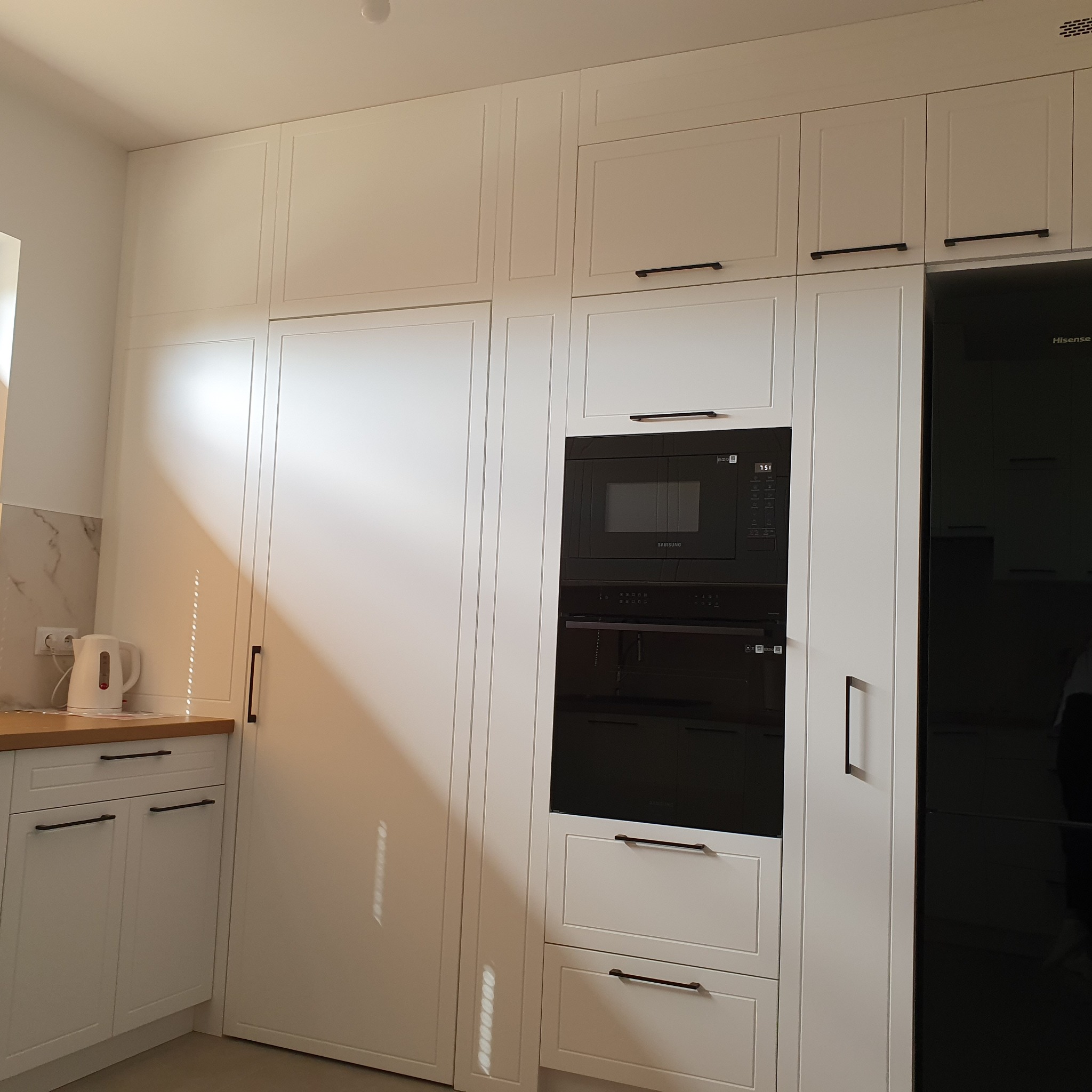 Biała kuchnia na wymiar z czarnymi uchwytami, zabudowanym sprzętem AGD (mikrofalówka i piekarnik) oraz lodówką. Nowoczesny minimalizm.