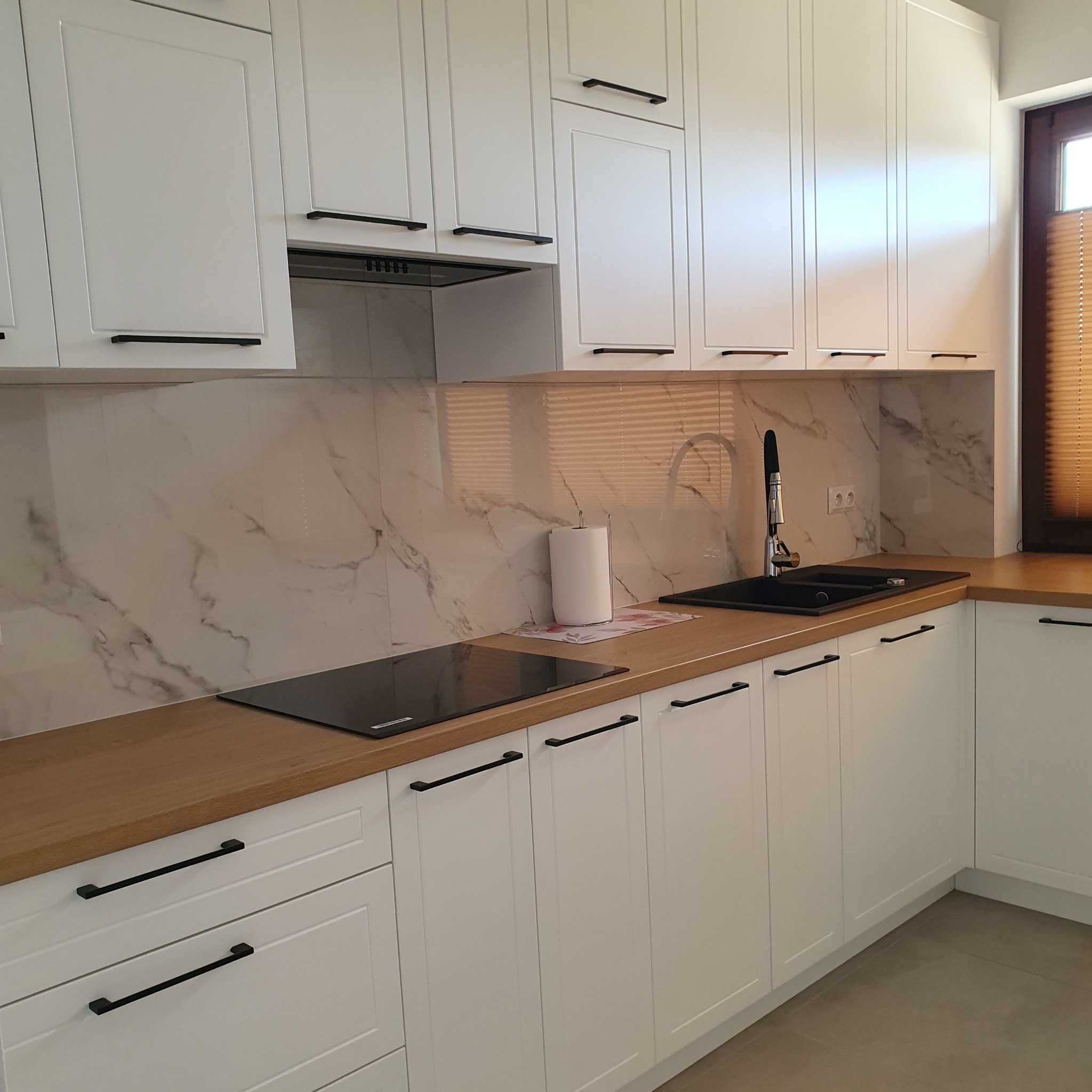 Nowoczesna, biała kuchnia z drewnianym blatem i czarnymi uchwytami. Marmurowe panele na ścianie. Widoczna płyta indukcyjna i zlew. Subtelne oświetlenie.