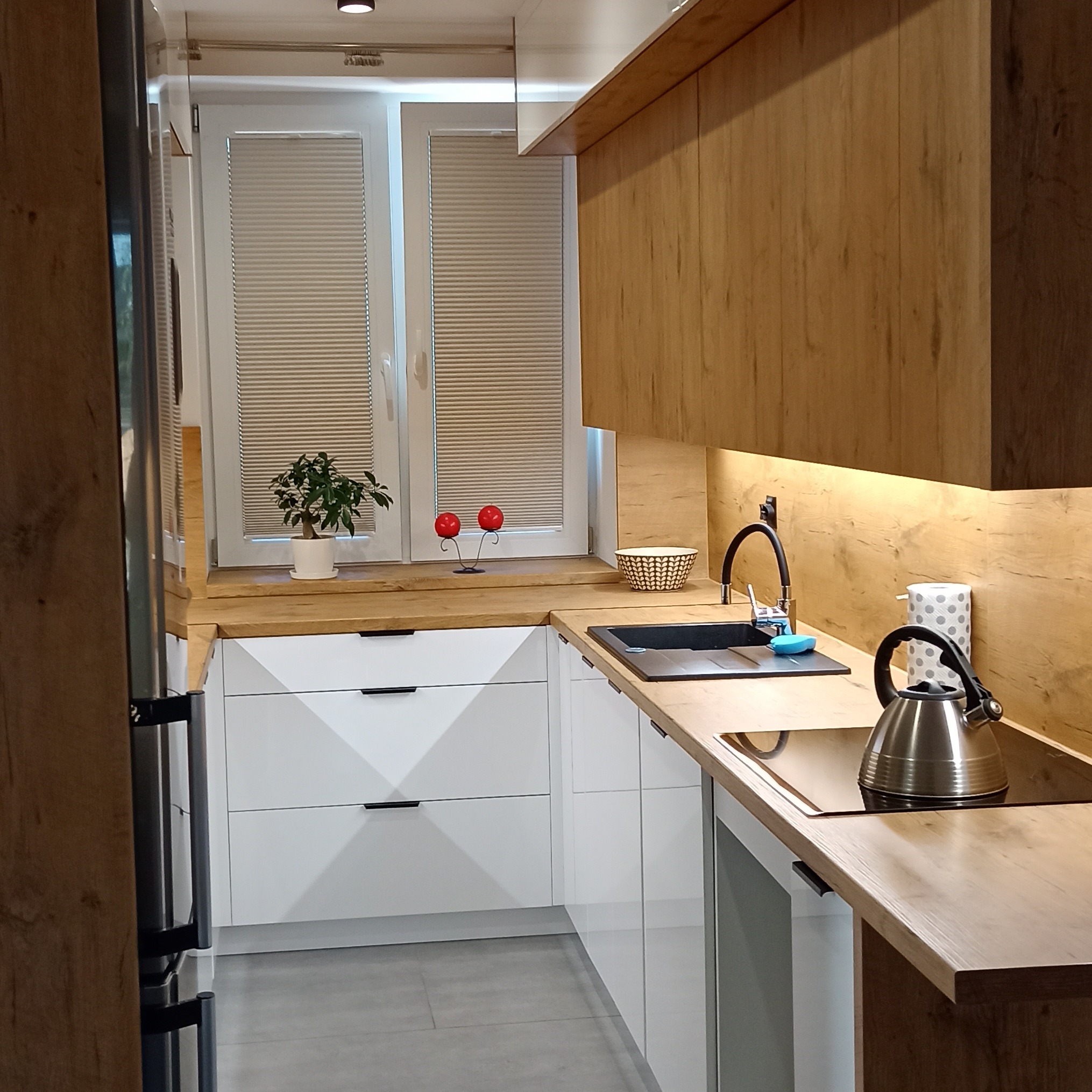 Nowoczesna kuchnia z białymi frontami i drewnianym blatem. Uwagę przykuwają minimalistyczne uchwyty i oświetlenie LED pod szafkami górnymi. Na blacie czajnik.