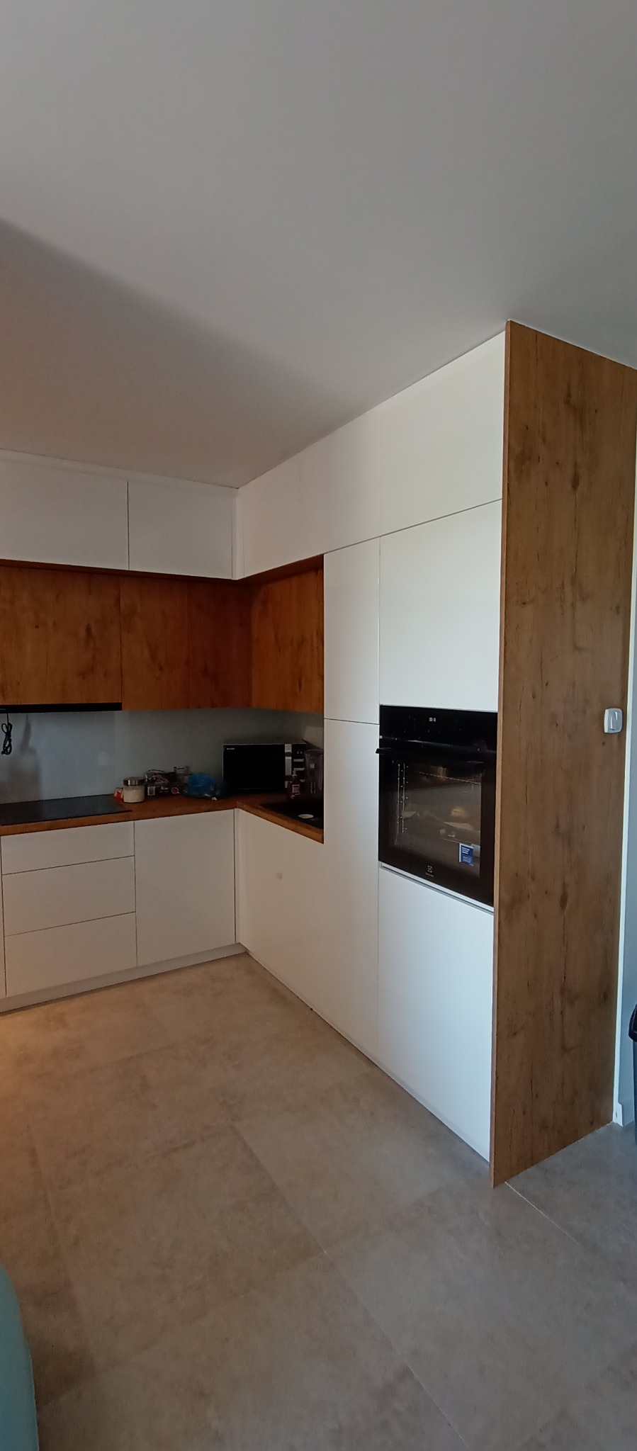 Nowoczesna kuchnia z białymi frontami i drewnianymi akcentami. Widoczny piekarnik w zabudowie i blat z kuchenką. Jasna podłoga. Ujęcie z perspektywy podłogi.