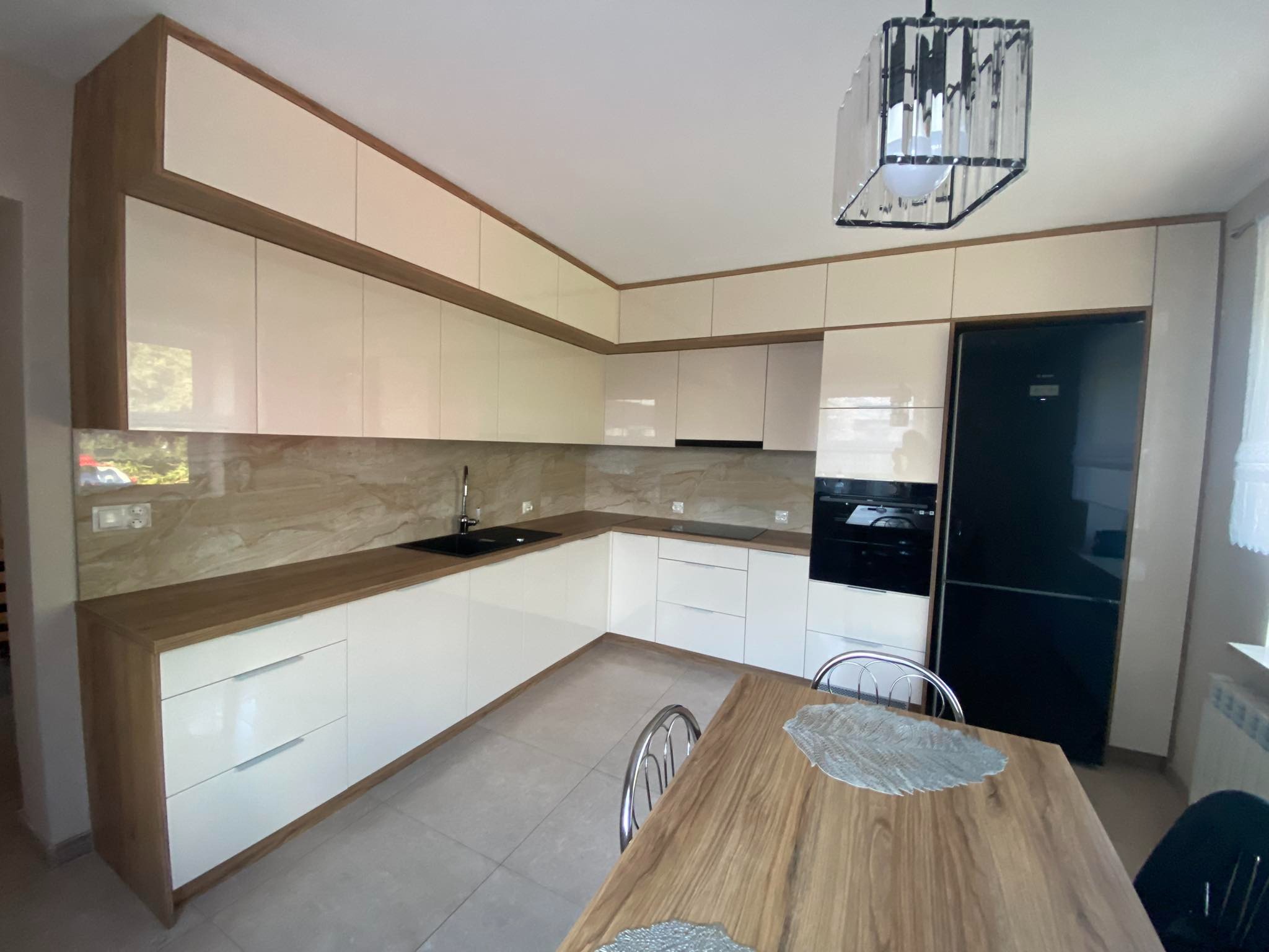 Jasna kuchnia na wymiar z drewnianymi akcentami, czarnym zlewem i lodówką. Nad stołem kryształowy żyrandol. Widok na całe pomieszczenie, ukończona realizacja.