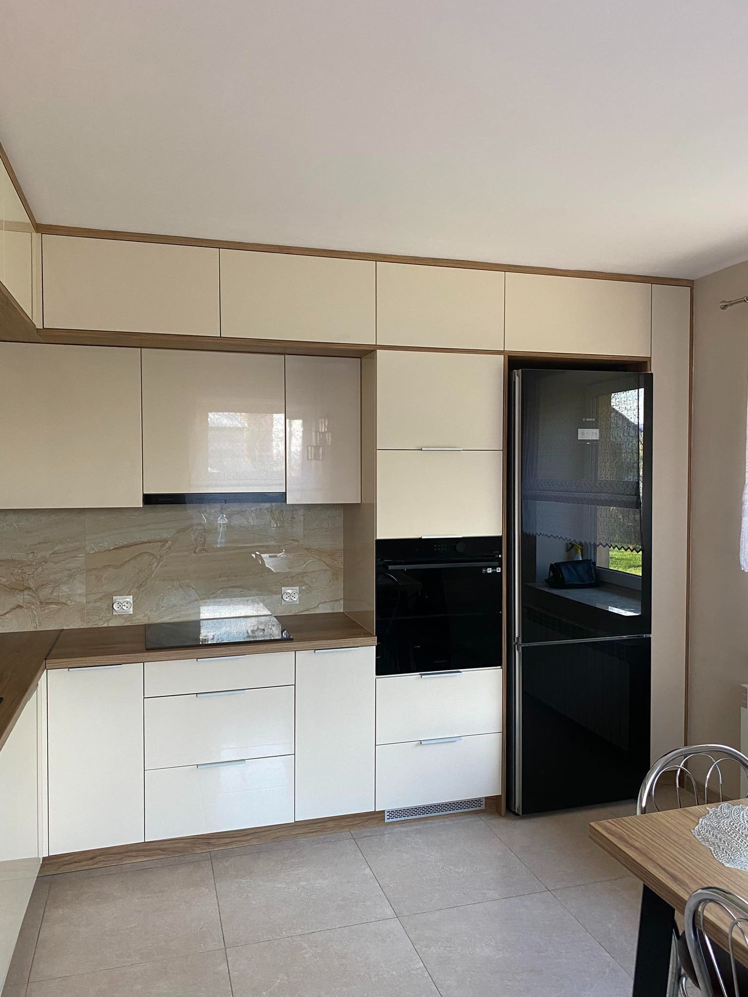 Jasna kuchnia z beżowymi frontami, drewnianymi akcentami i czarną lodówką Bosch, widok na płytki imitujące marmur na ścianie.