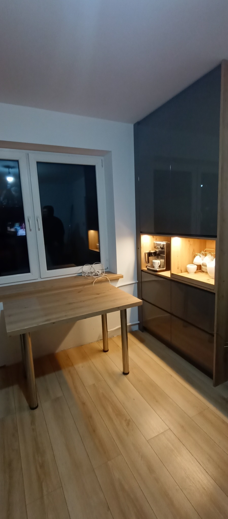 Nowoczesna zabudowa kuchenna z szarymi frontami i drewnianym blatem w Bielawach. Wnęka z oświetleniem eksponuje ekspres do kawy i filiżanki.