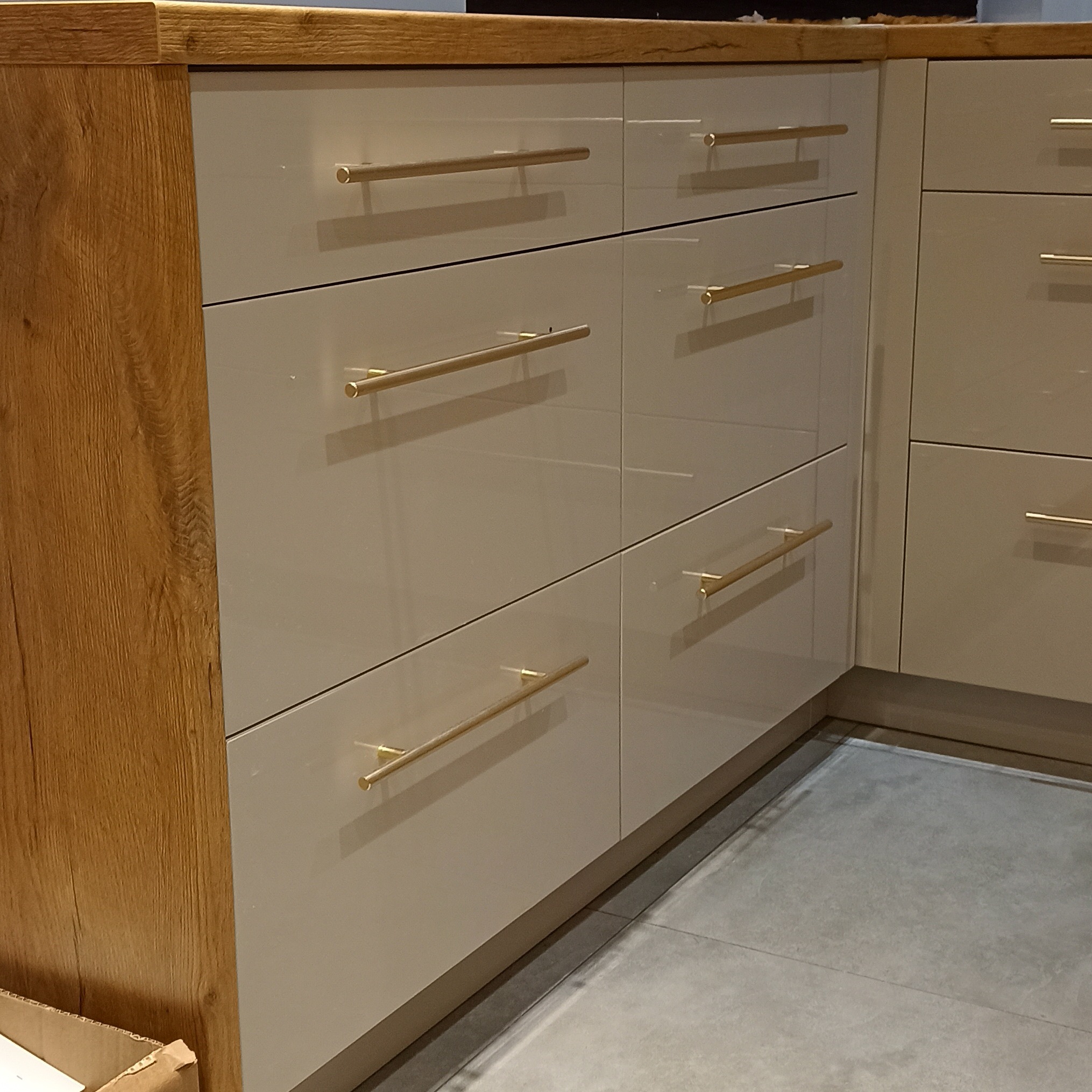 Beżowe, lakierowane fronty szuflad z podłużnymi, złotymi uchwytami, drewniany blat i bok szafki w kuchni na wymiar. Widoczny fragment szarej podłogi.