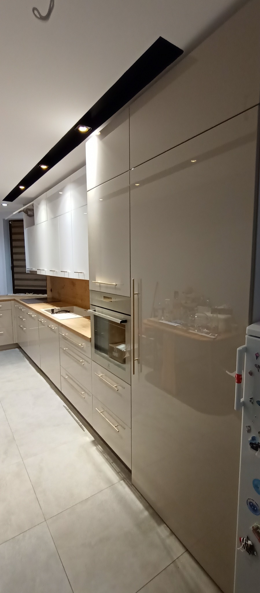 Jasna kuchnia z połyskiem, czarny pas oświetleniowy pod sufitem. Widoczne fronty szafek, blat z drewna, piekarnik i lodówka z magnesami. Minimalistyczny design.