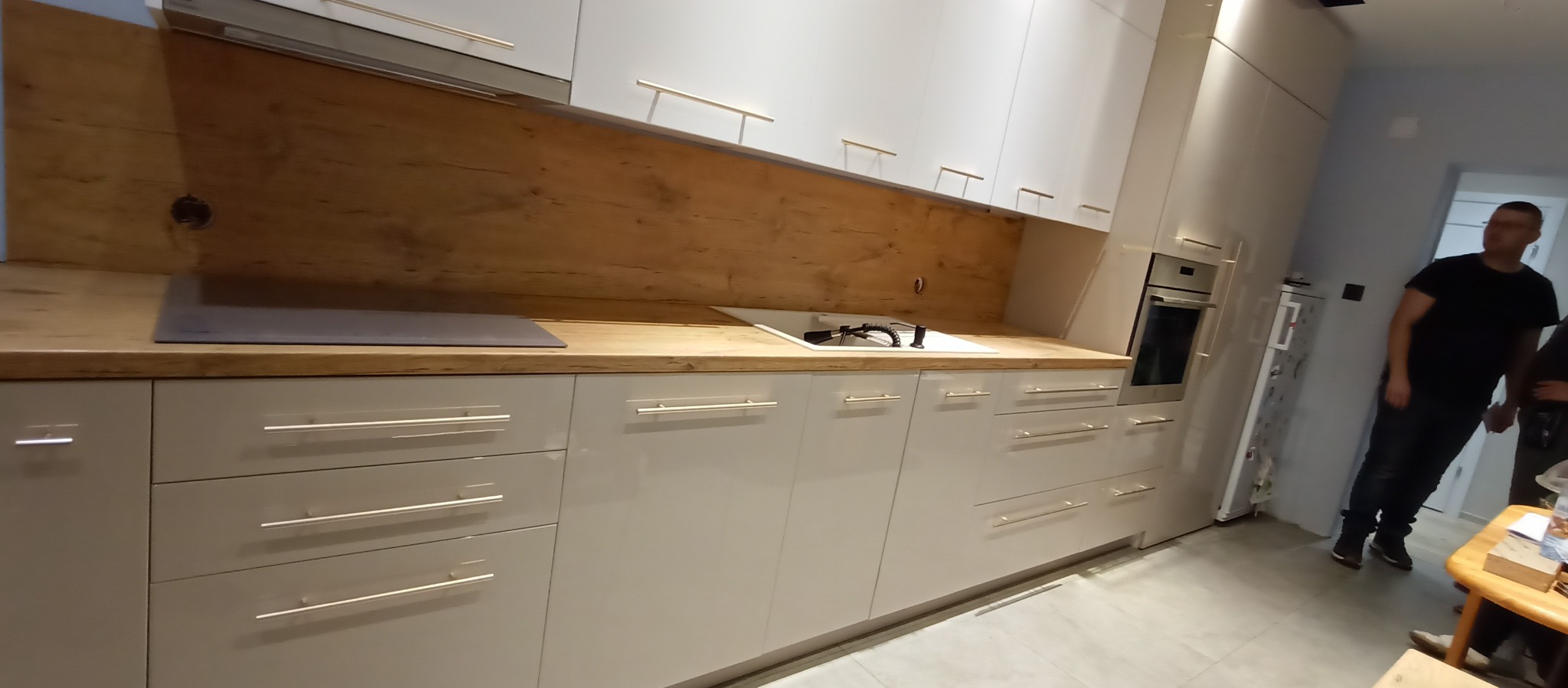 Jasna kuchnia na wymiar z drewnianym blatem i frontami z podłużnymi uchwytami, widoczny fragment płyty grzewczej i zlewu. Na drugim planie osoby.