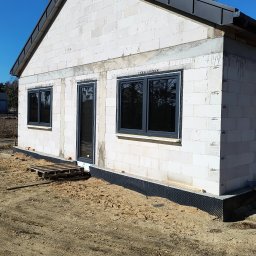 Dach-Expert Igor Smirnow - Surowy stan domu jednorodzinnego z szarego bloczka, z zamontowanymi oknami i drzwiami, oraz częściowo wykonanym dachem z blachy w kolorze grafitowym.