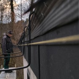 Dach-Expert Igor Smirnow - Montaż haków na dachu z blachy trapezowej przez dekarza na rusztowaniu. Widok z dołu, z żółtą linką poziomującą. W tle drzewa bez liści.