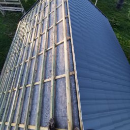 Dach-Expert Igor Smirnow - Częściowo pokryty dach z widoczną konstrukcją drewnianą i membraną. Fragment dachu pokryty ciemną blachą. Widok z góry na osobę stojącą na dachu w trakcie prac.