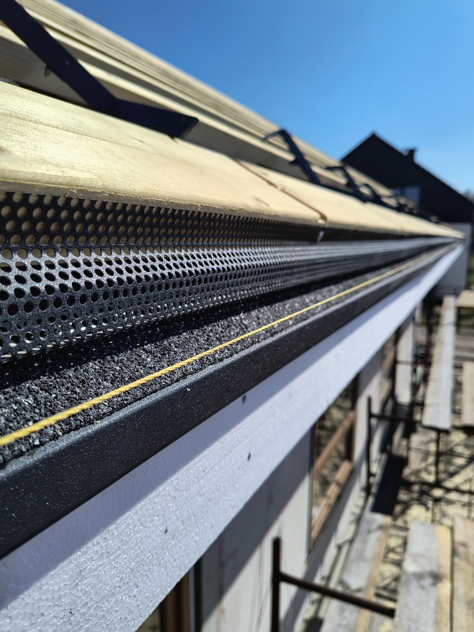 Zamontowana siatka zabezpieczająca rynnę przed liśćmi i innymi zanieczyszczeniami. Widoczne drewniane elementy konstrukcji dachu i ocieplenie budynku. Ujęcie z dołu, w słoneczny dzień.