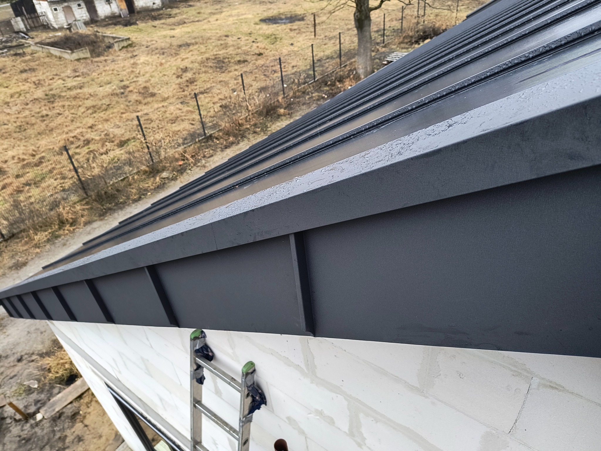 Nowe, grafitowe pokrycie dachowe z blachy na budynku w Żyrardowie, widok z góry. Na ścianie oparta drabina. Widoczne krople wody na powierzchni.