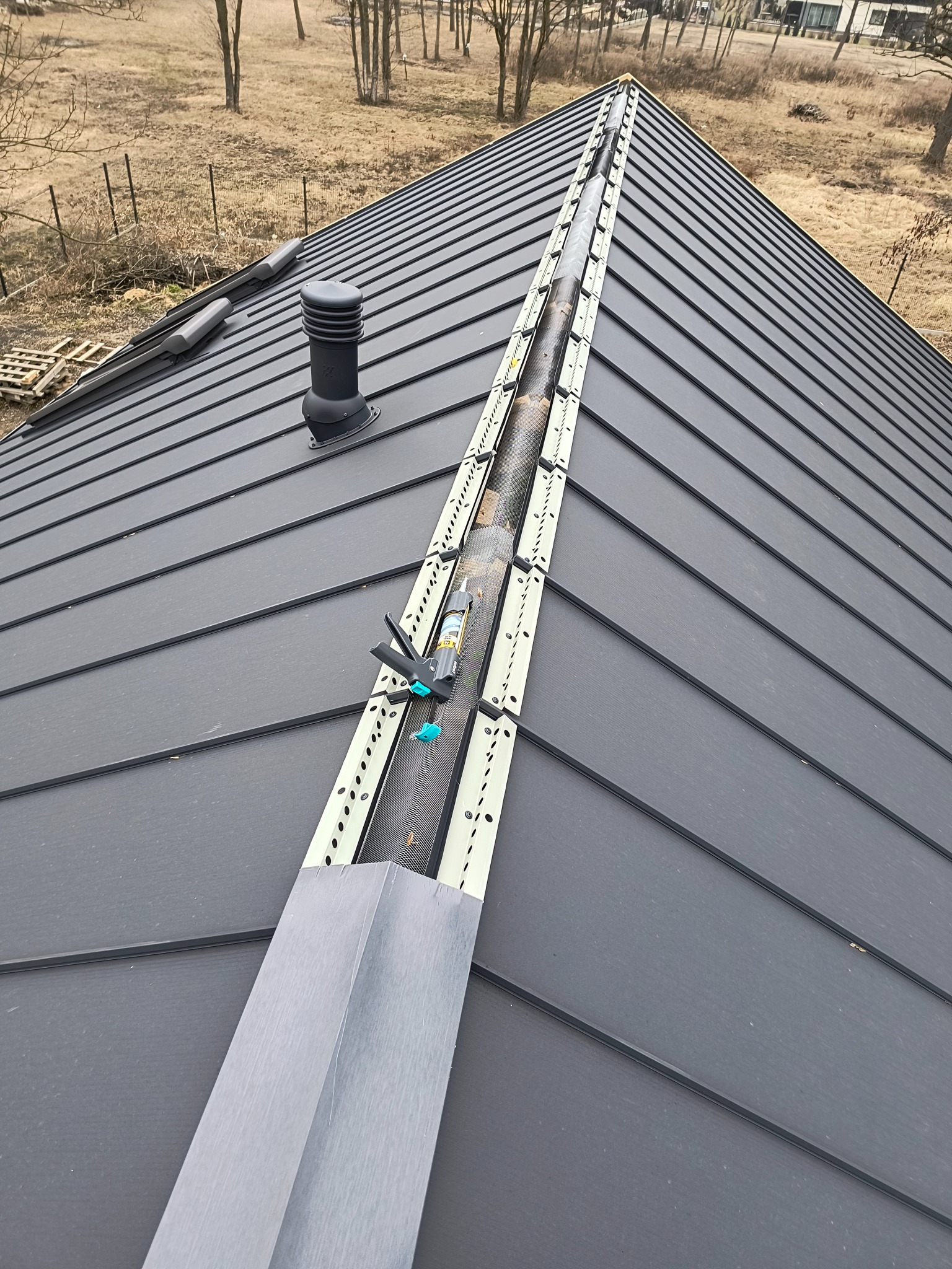 Montaż systemu rynnowego na grafitowym dachu z blachy na rąbek stojący. Widoczny kominek wentylacyjny i narzędzia montażowe. Ujęcie z góry, w tle drzewa.