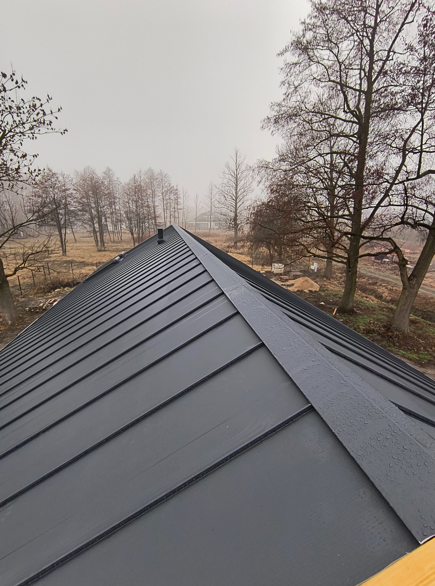Szary dach z blachy na tle drzew w mglisty dzień. Widoczne krople wody na powierzchni. Ujęcie z góry, podkreślające geometrię i fakturę pokrycia.