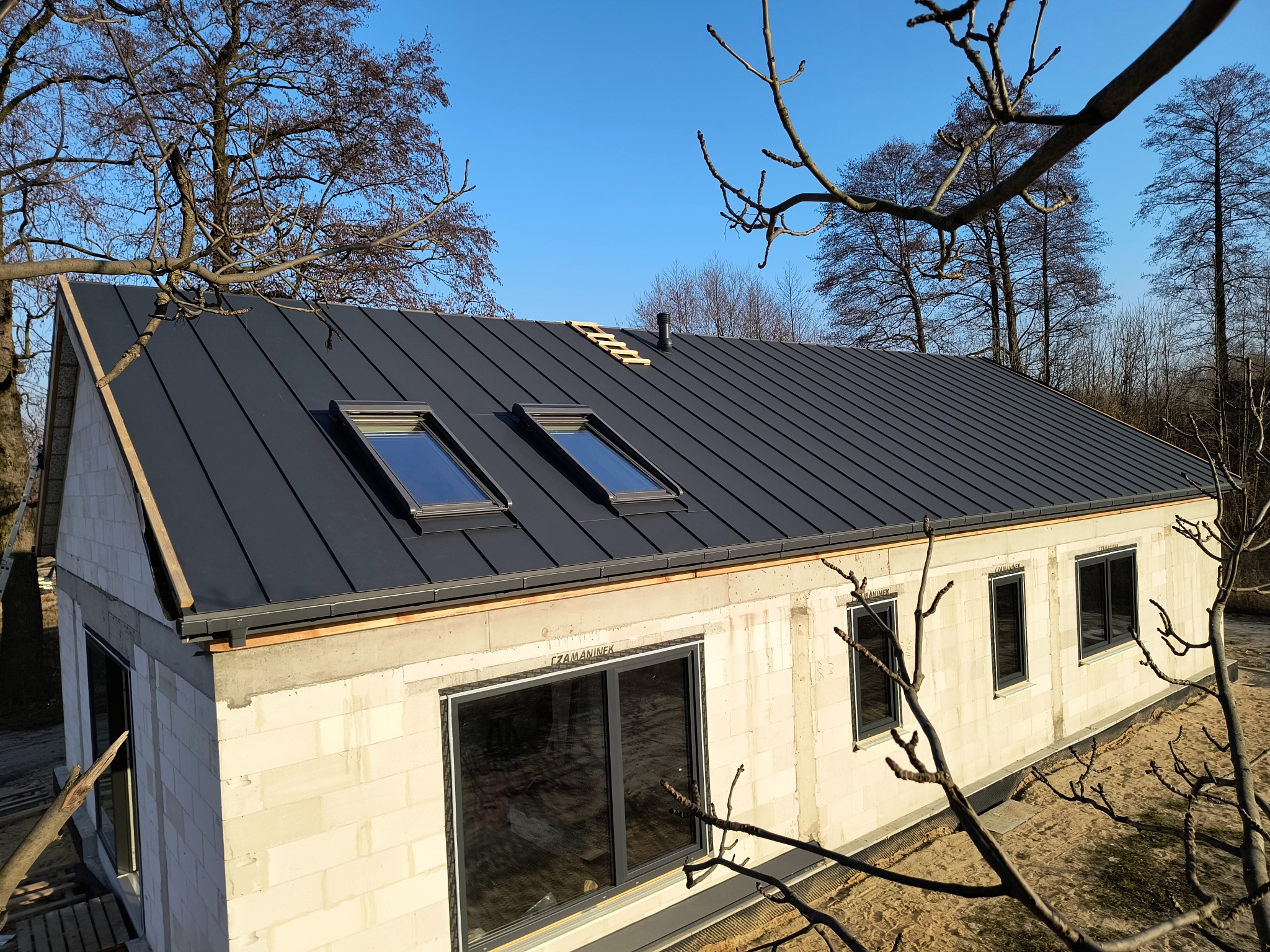 Dom w stanie surowym zamkniętym z ciemnym dachem z blachy na rąbek stojący i oknami dachowymi. Widok z góry przez gałęzie drzew.